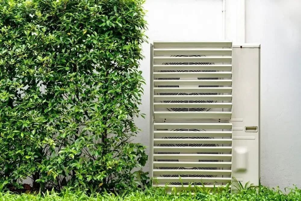 A White Air Conditioning Unit is Installed Against a Wall, Partially Shaded by a Lush Green Bush 
