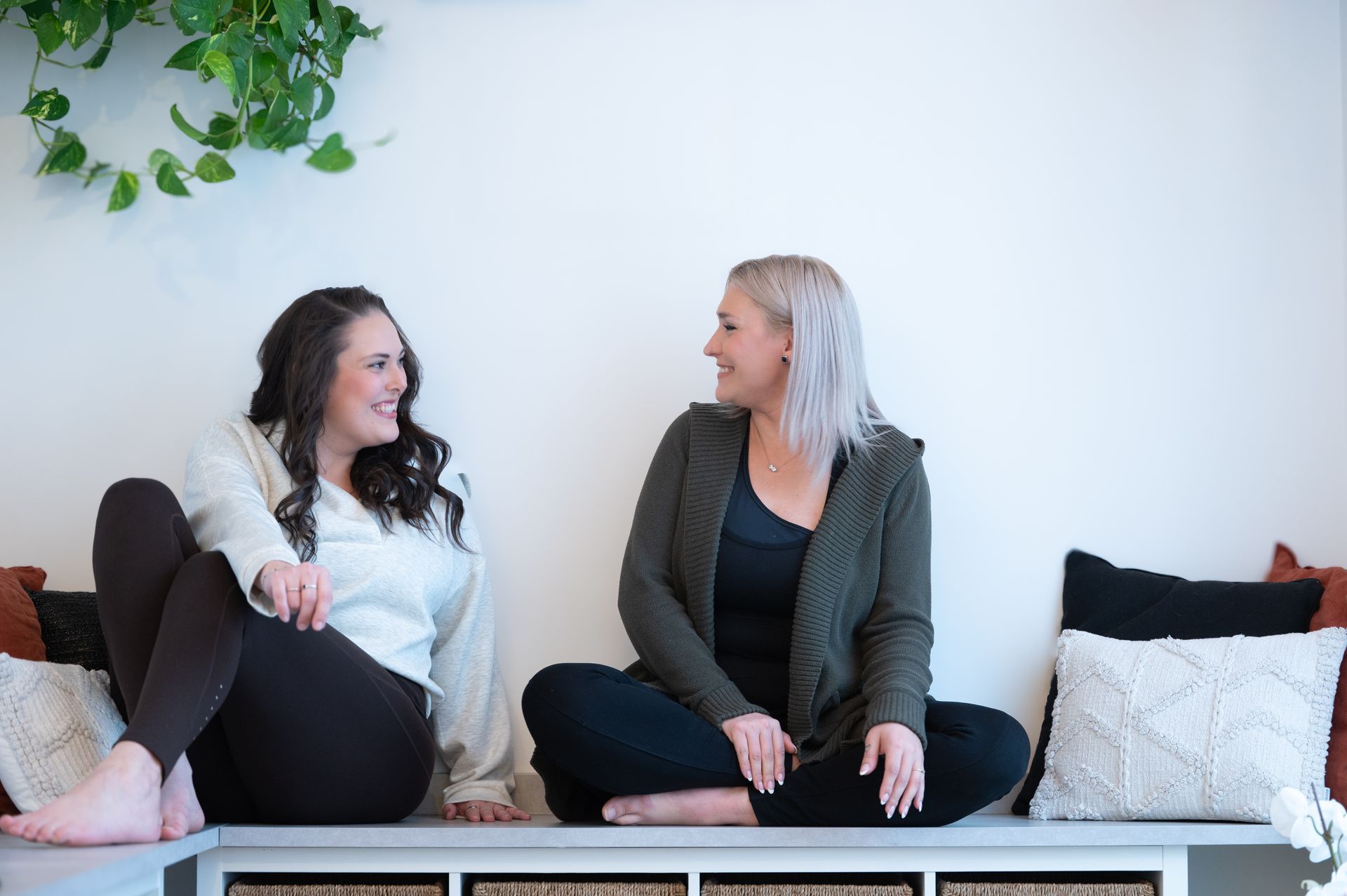 Deux femmes assises sur un banc se sourient. Mur blanc, verdure, coussins.