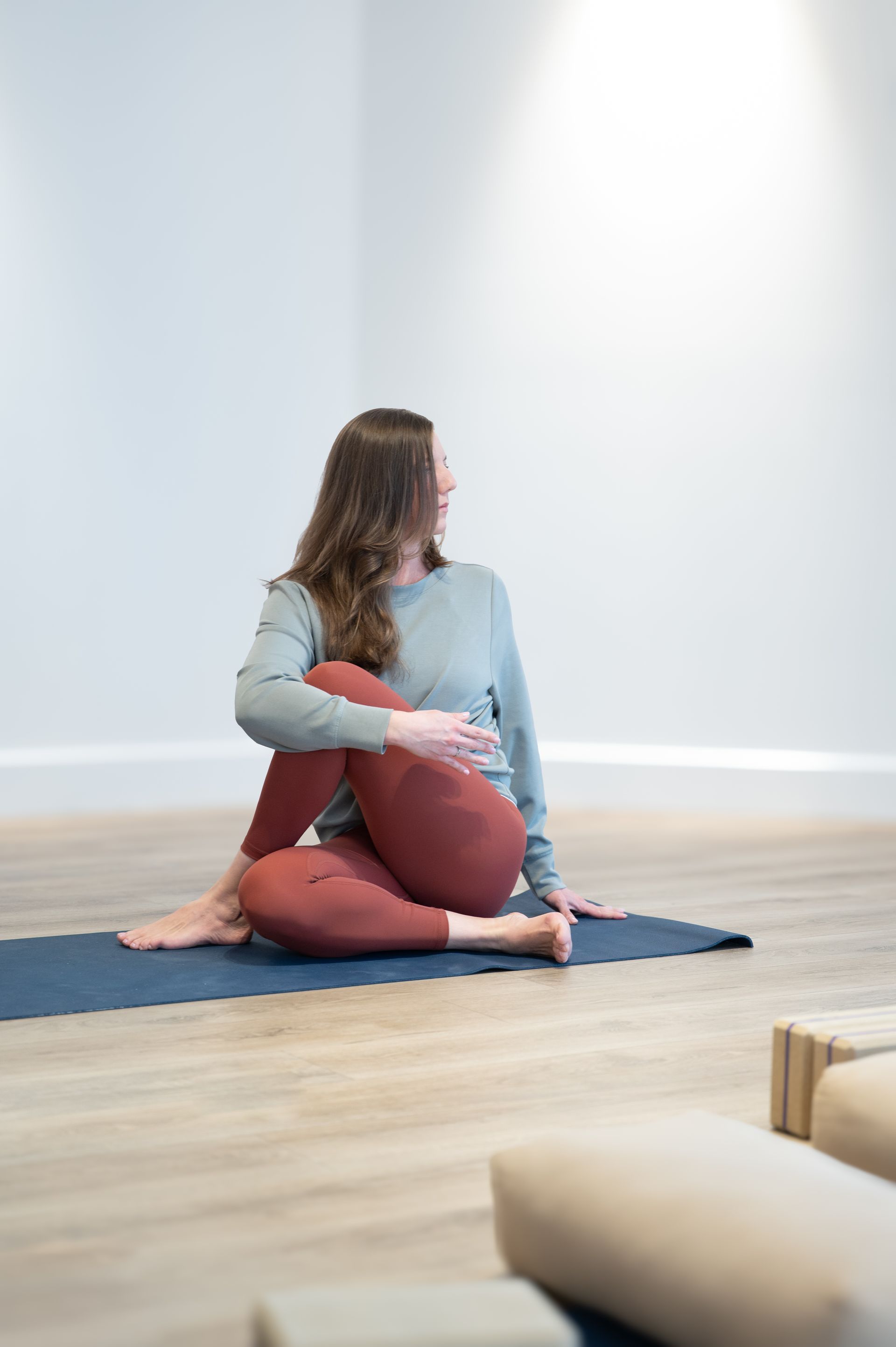 Femme dans une pose de yoga en torsion assise sur un tapis bleu, portant des leggings rouges et un sweat-shirt gris, dans un studio.