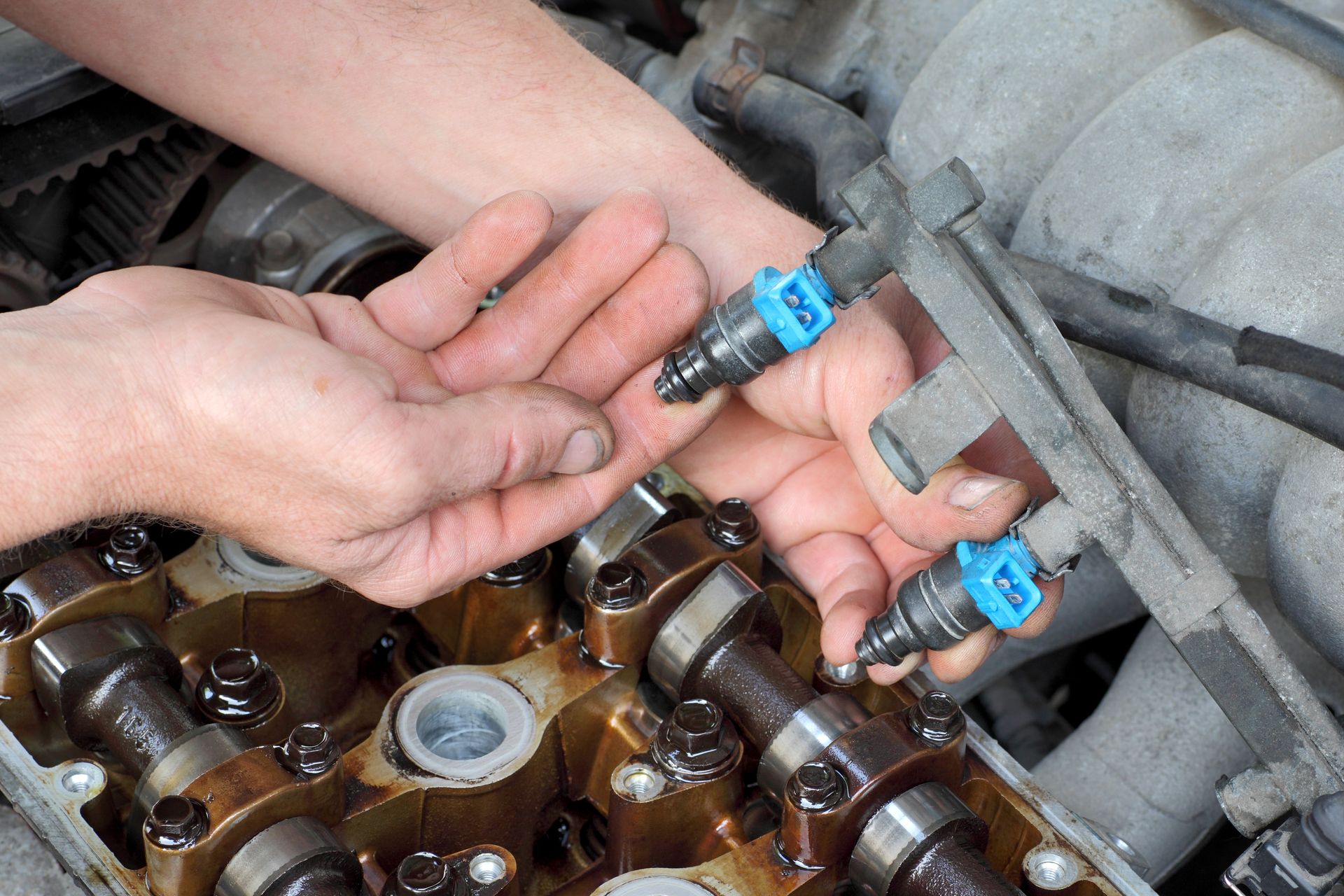 Hands holding blue automotive fuel injectors attached to a metal fuel rail, positioned over an open car engine.