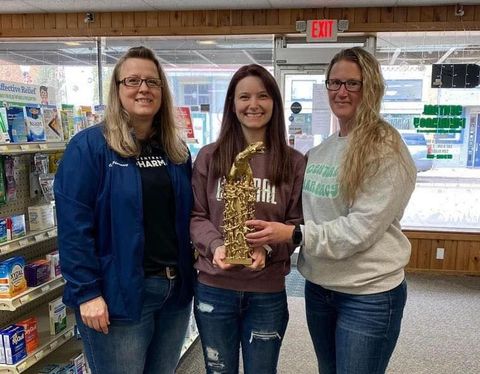 Three women are standing next to each other in a pharmacy holding a trophy.