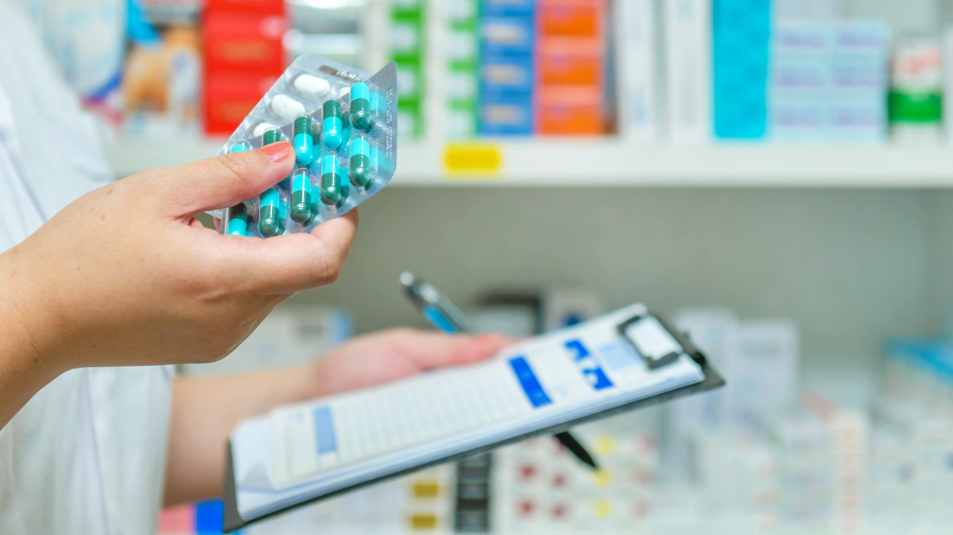 A pharmacist is holding a blister pack of pills and writing on a clipboard.