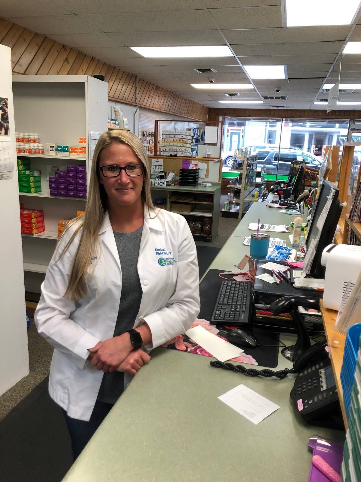 A woman in a lab coat is standing at a counter in a pharmacy.