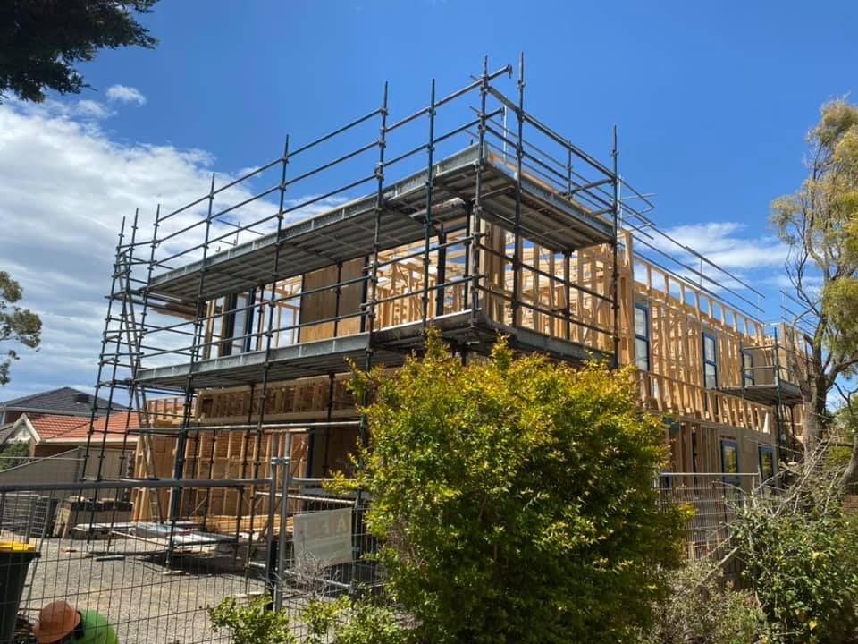 Two-story house under construction with scaffolding. Wooden frame visible. Blue sky, green bushes in front.