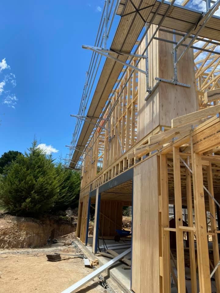 Construction site: wooden frame of a building with scaffolding, blue sky, and green trees.