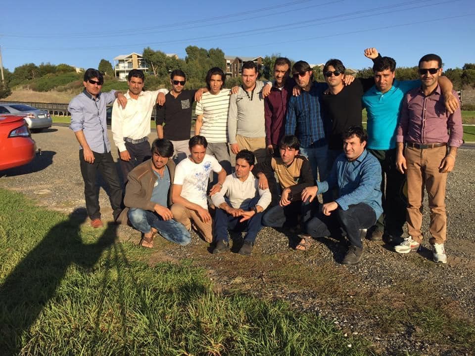 Group of men standing and kneeling on gravel, smiling, with blue sky and trees in the background.