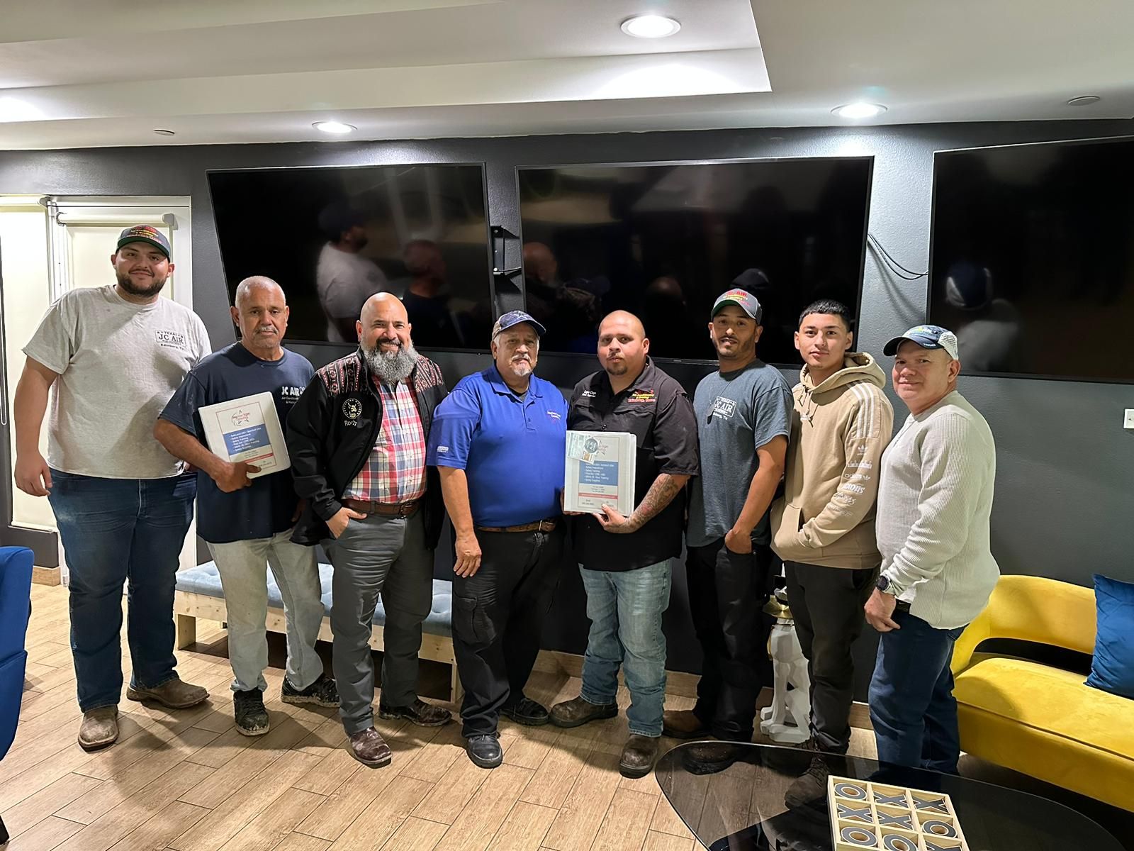 A group of men, some holding papers, pose in front of large screens. Inside a room with couches.