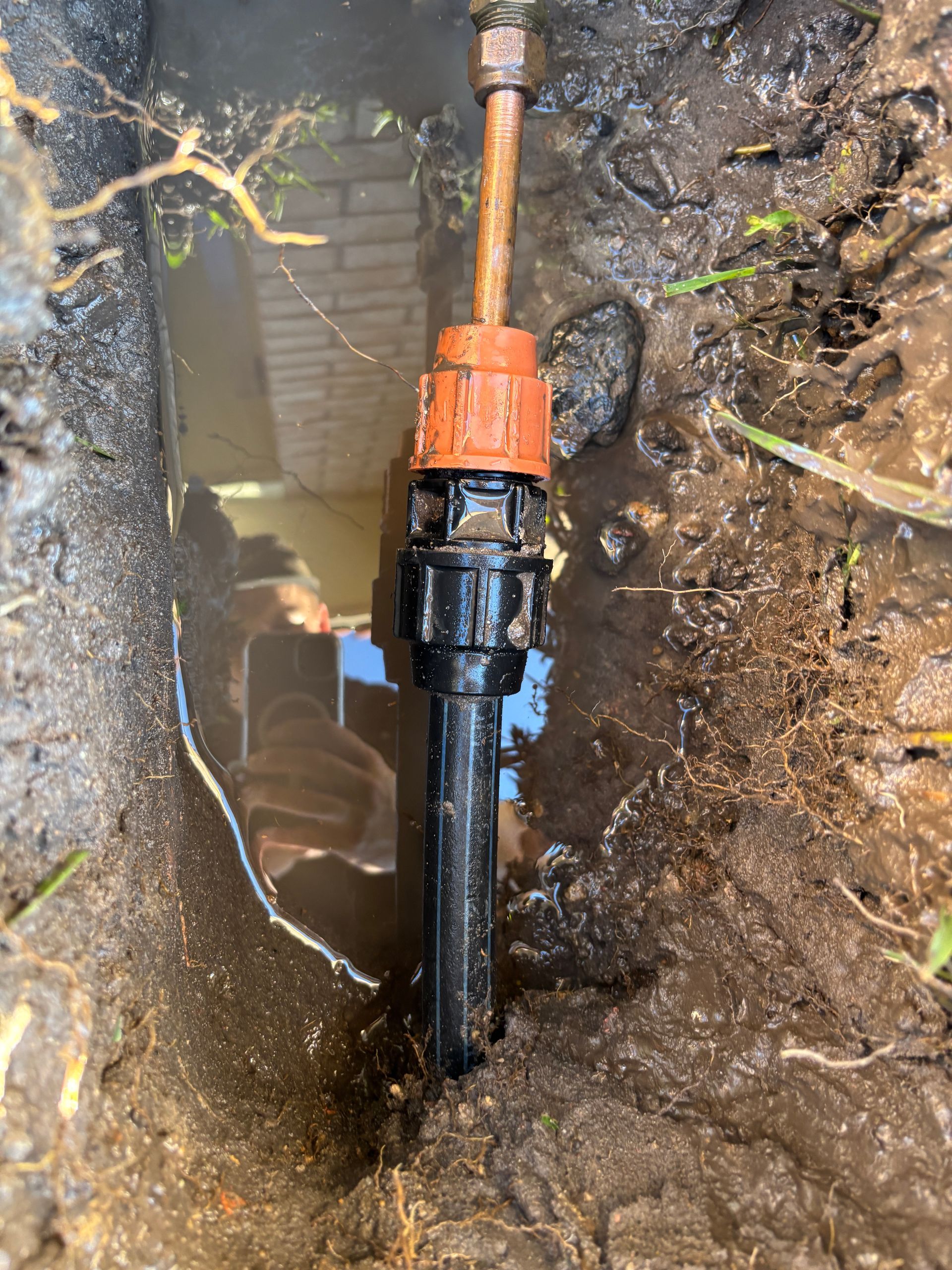 Black sprinkler head in muddy trench, with copper pipe and orange fitting.