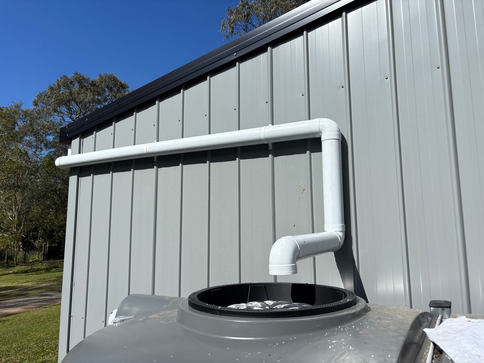 White pipes divert rainwater from a metal shed's gutter into a gray water tank. Sunny day.