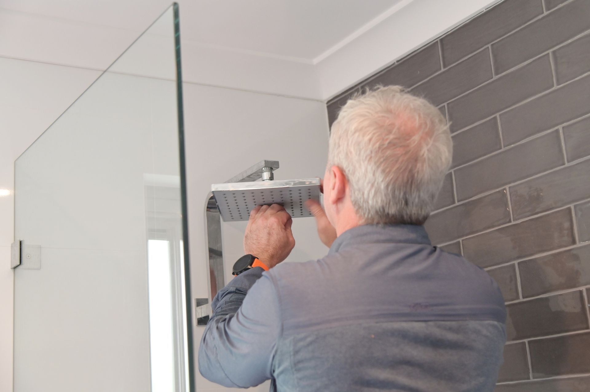 Man installs a square, chrome showerhead in a bathroom with gray tile.