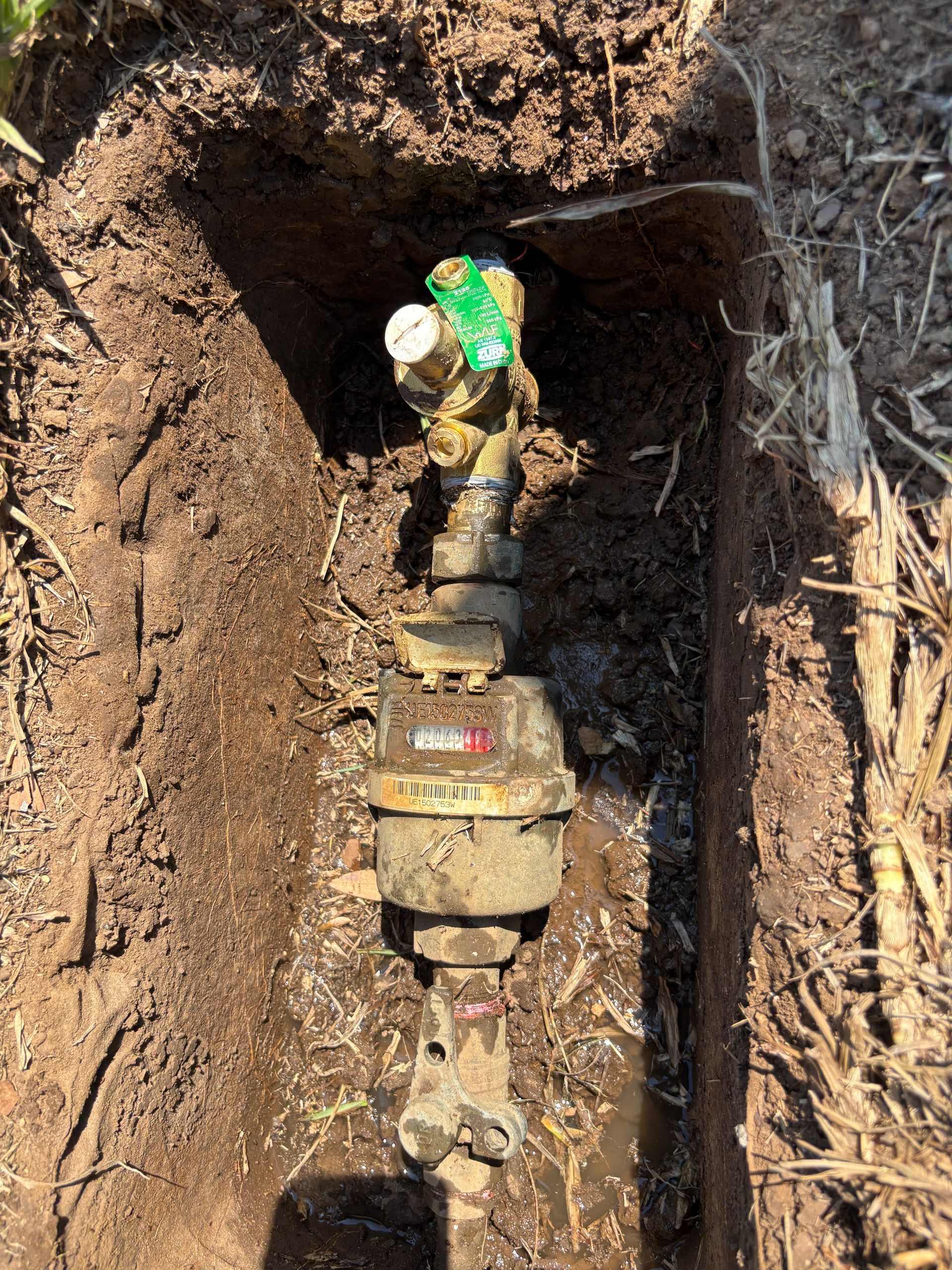 A water meter in a muddy trench, with pipes and a gauge visible.