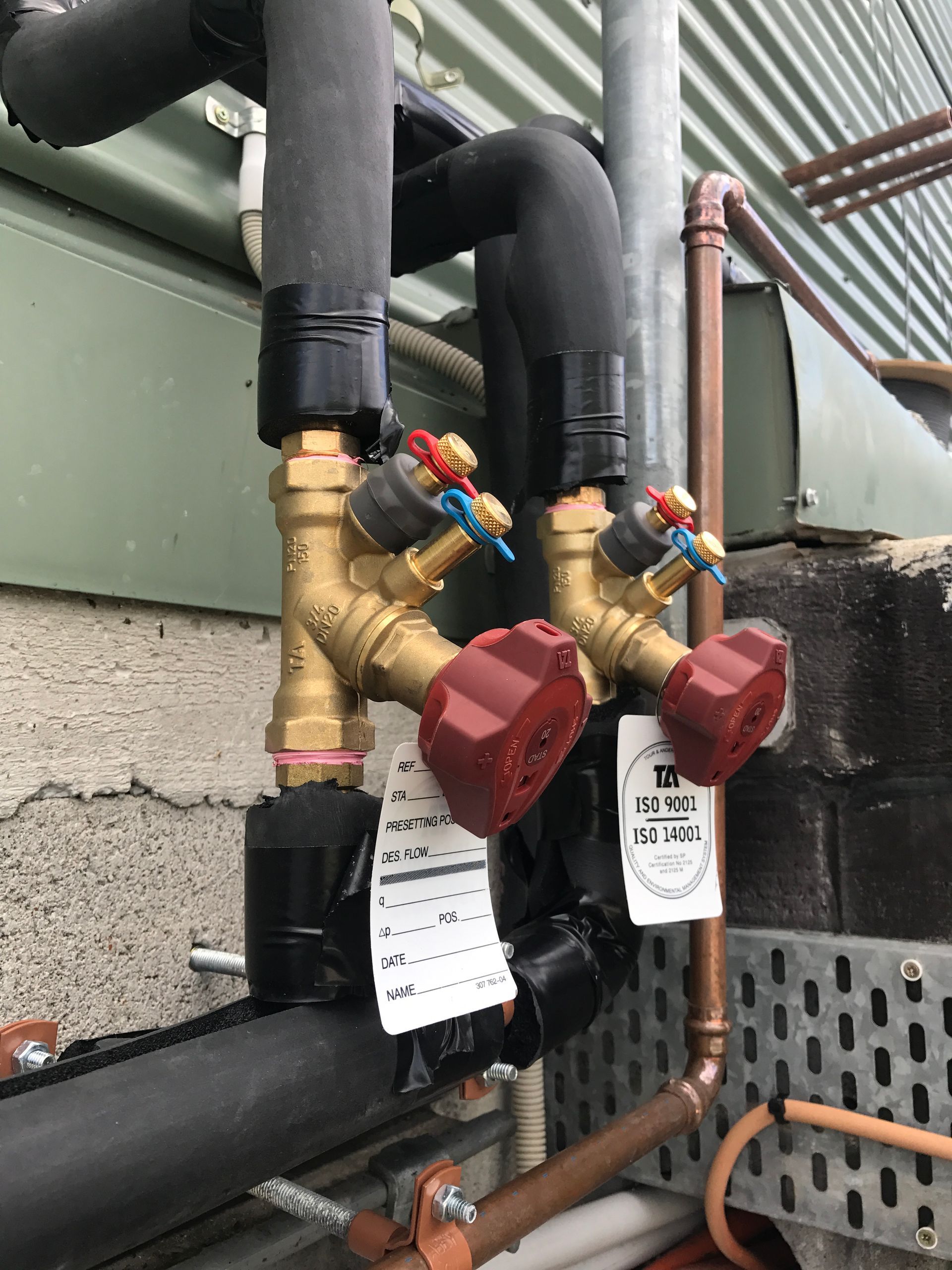 Pipes with red and blue valves and black insulation attached to a building's exterior.