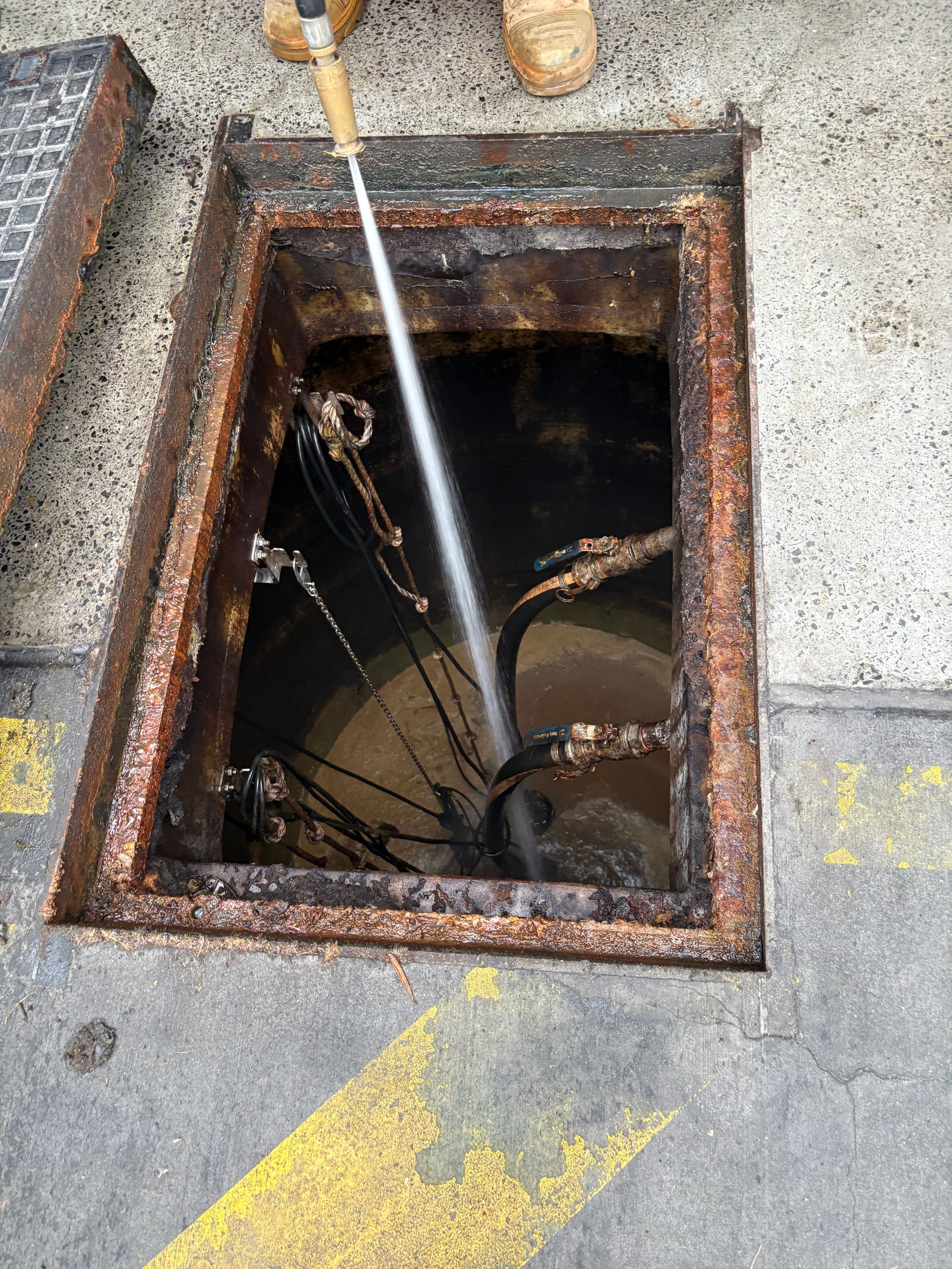 A person power washes inside an open, rusty utility pit, sending water downward.