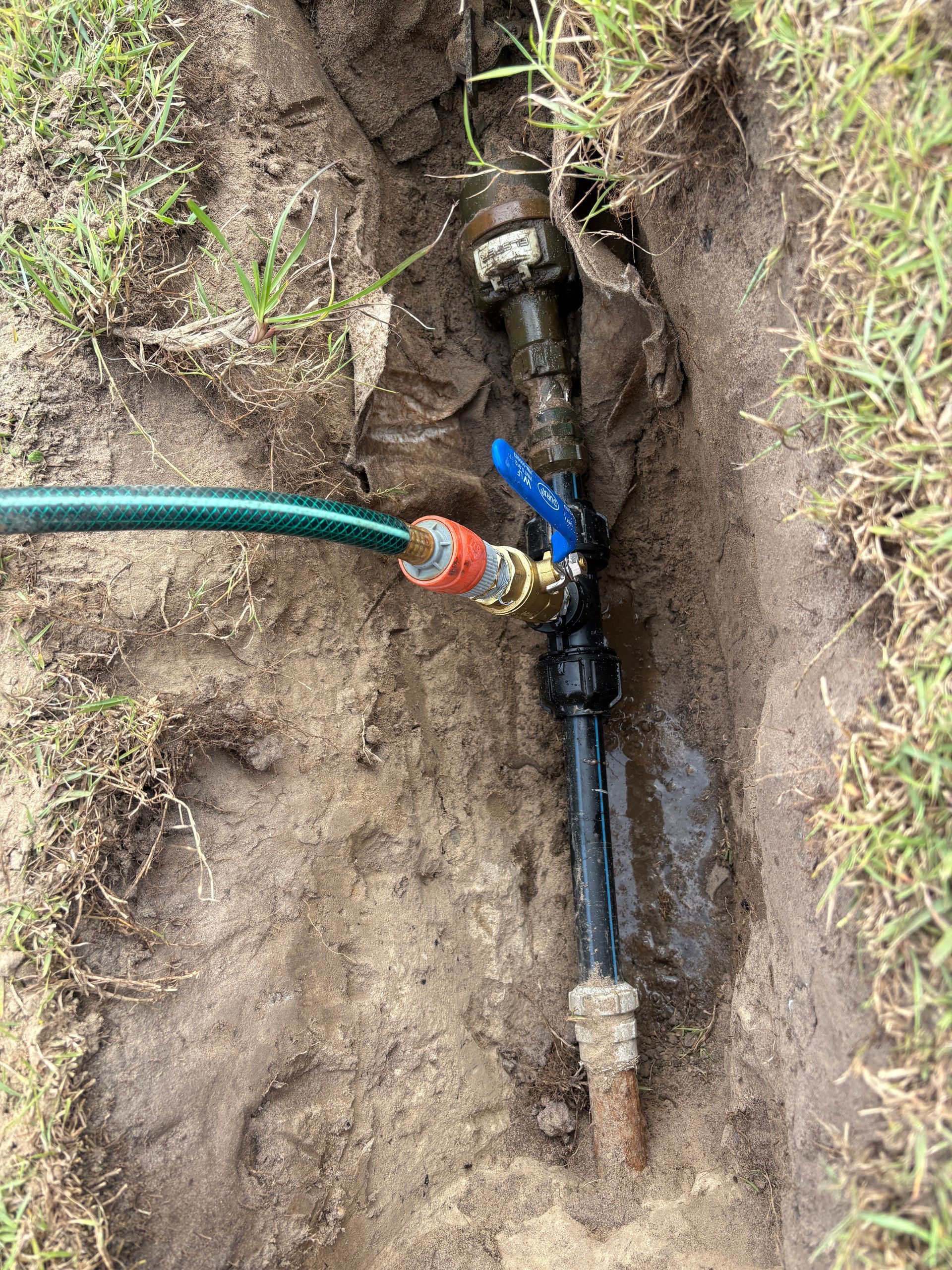 Water pipe with hose and valve exposed in a dug trench, surrounded by grass and soil.