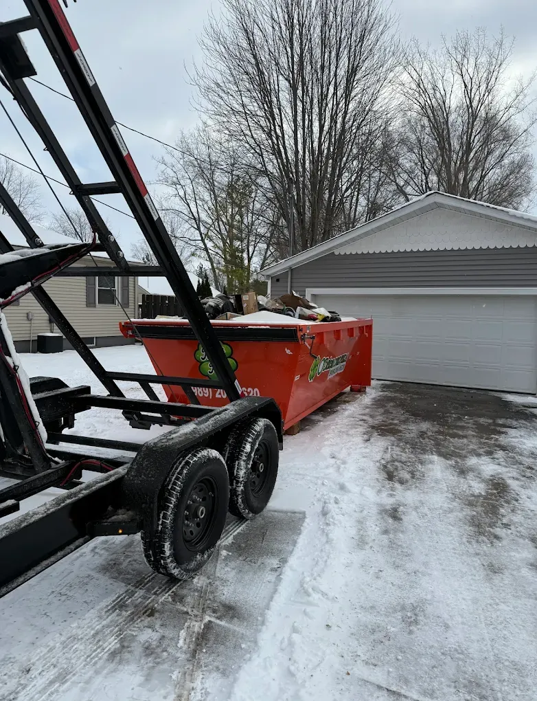 Orange dumpster on a trailer in snowy driveway, in front of a gray garage.
