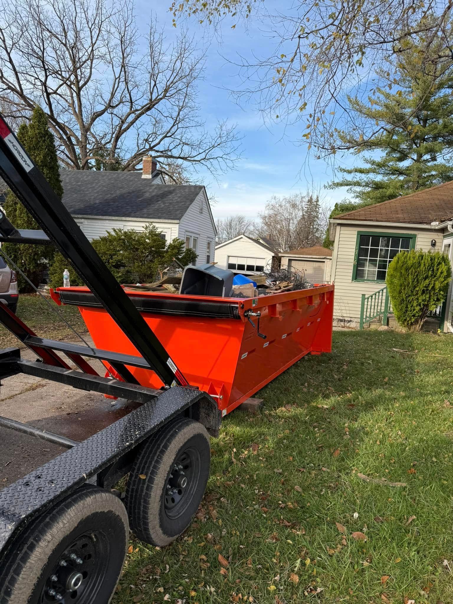 Orange debris container on trailer in a yard with houses and trees under a sunny sky.