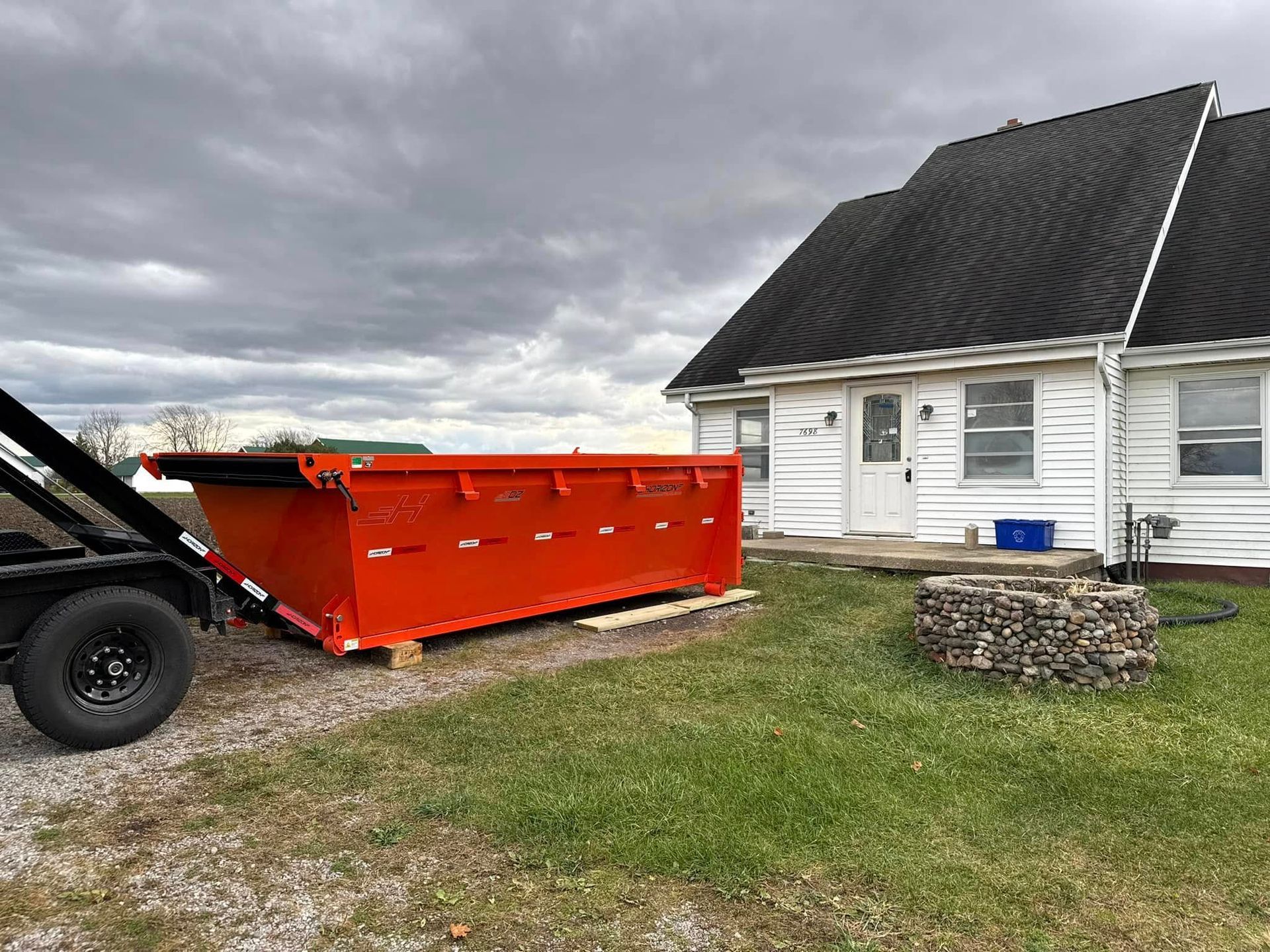 An orange dumpster on a trailer sits on a gravel driveway next to a white house with a dark roof on a cloudy day.