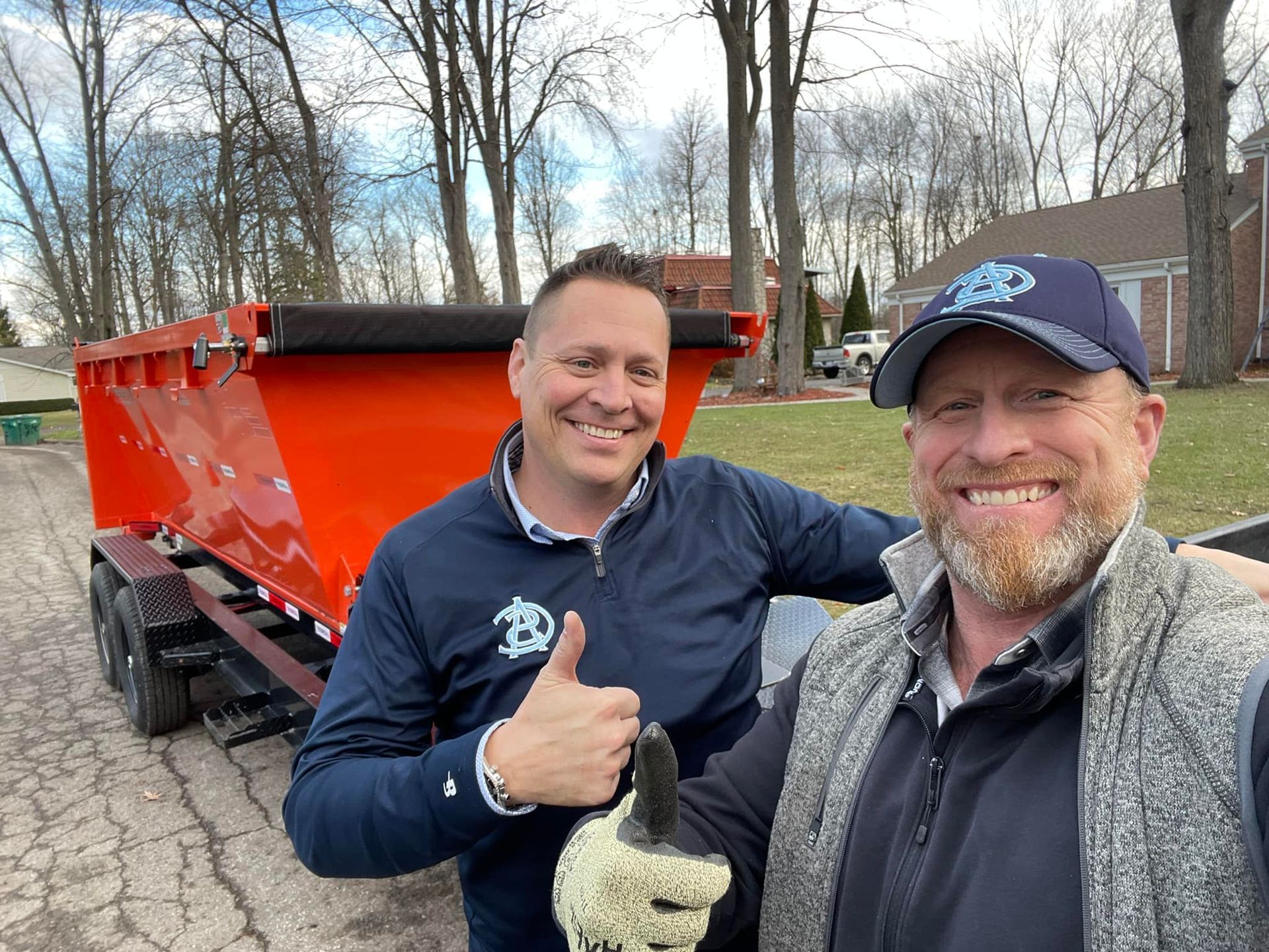 Two men smile next to an orange dump trailer. One gives a thumbs up. Outdoors, trees in background.