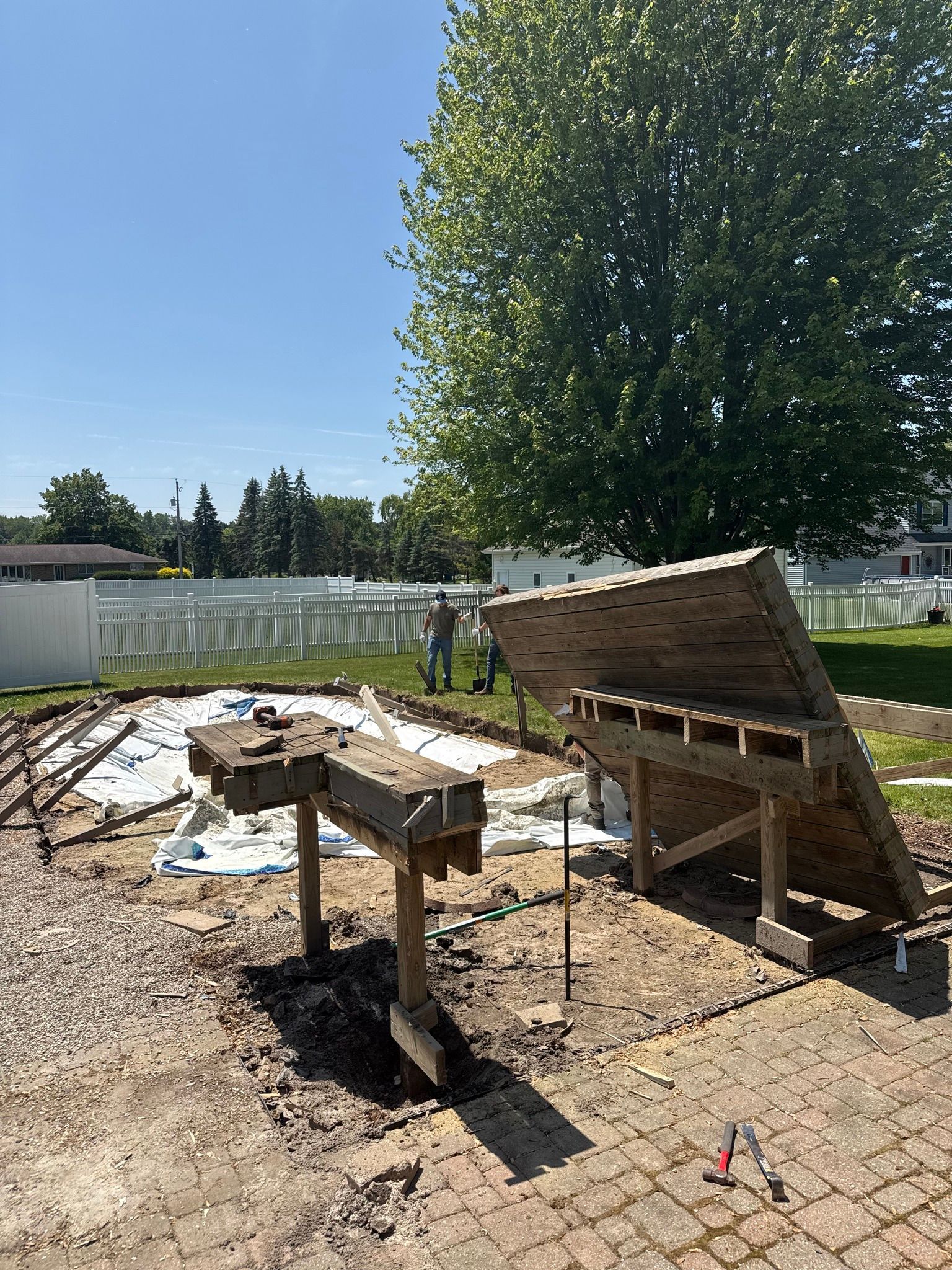 Construction site with wooden structures, ground partially excavated, person visible near a fence, sunny day.