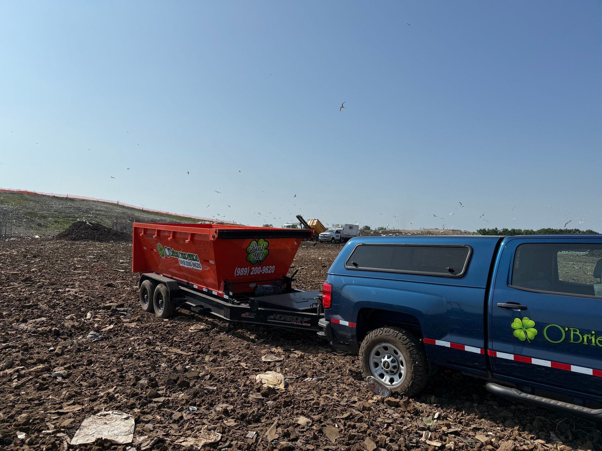 Blue truck towing an orange dumpster trailer in a landfill; sunny, clear sky.