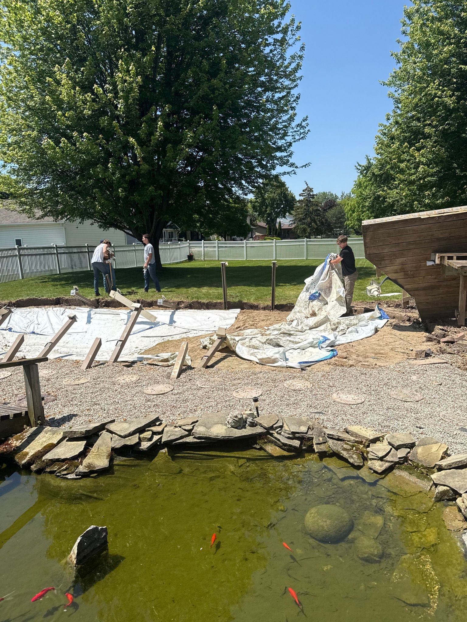 Workers constructing a patio by a pond with fish, using stones and sand under a sunny sky.