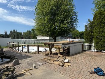 Pool under construction. Wooden frame and deck, with debris on a paved area, green trees, and a white fence.
