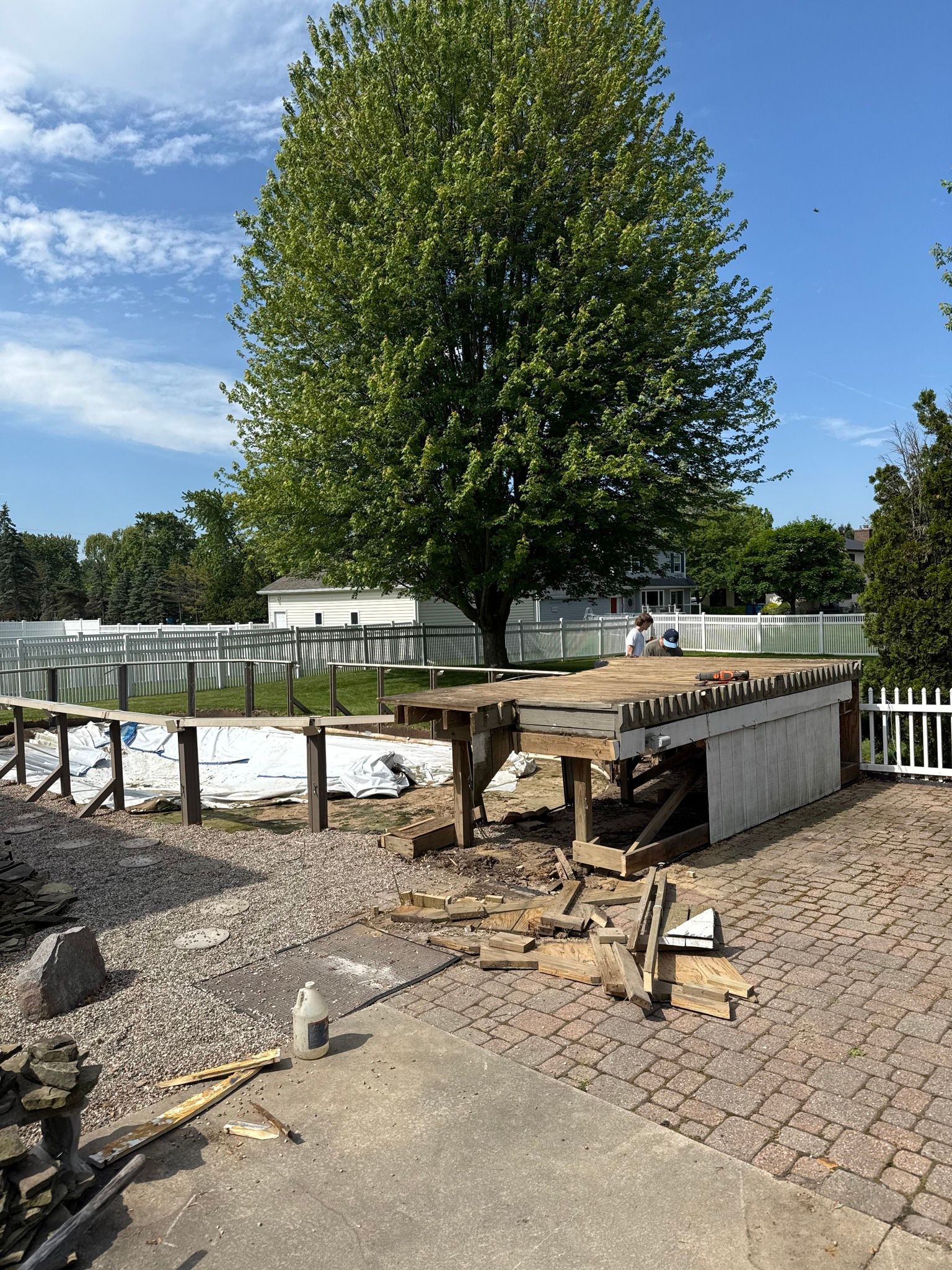 Construction site: a wooden structure being demolished next to a paved patio, large tree, and a fence.