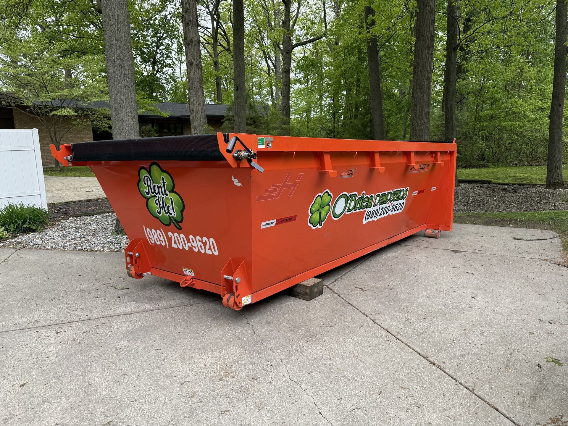 Orange dumpster on concrete, in front of trees. Green logo on side.