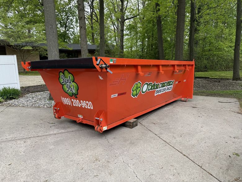 Orange dumpster on concrete, in front of trees. Green logo on side.