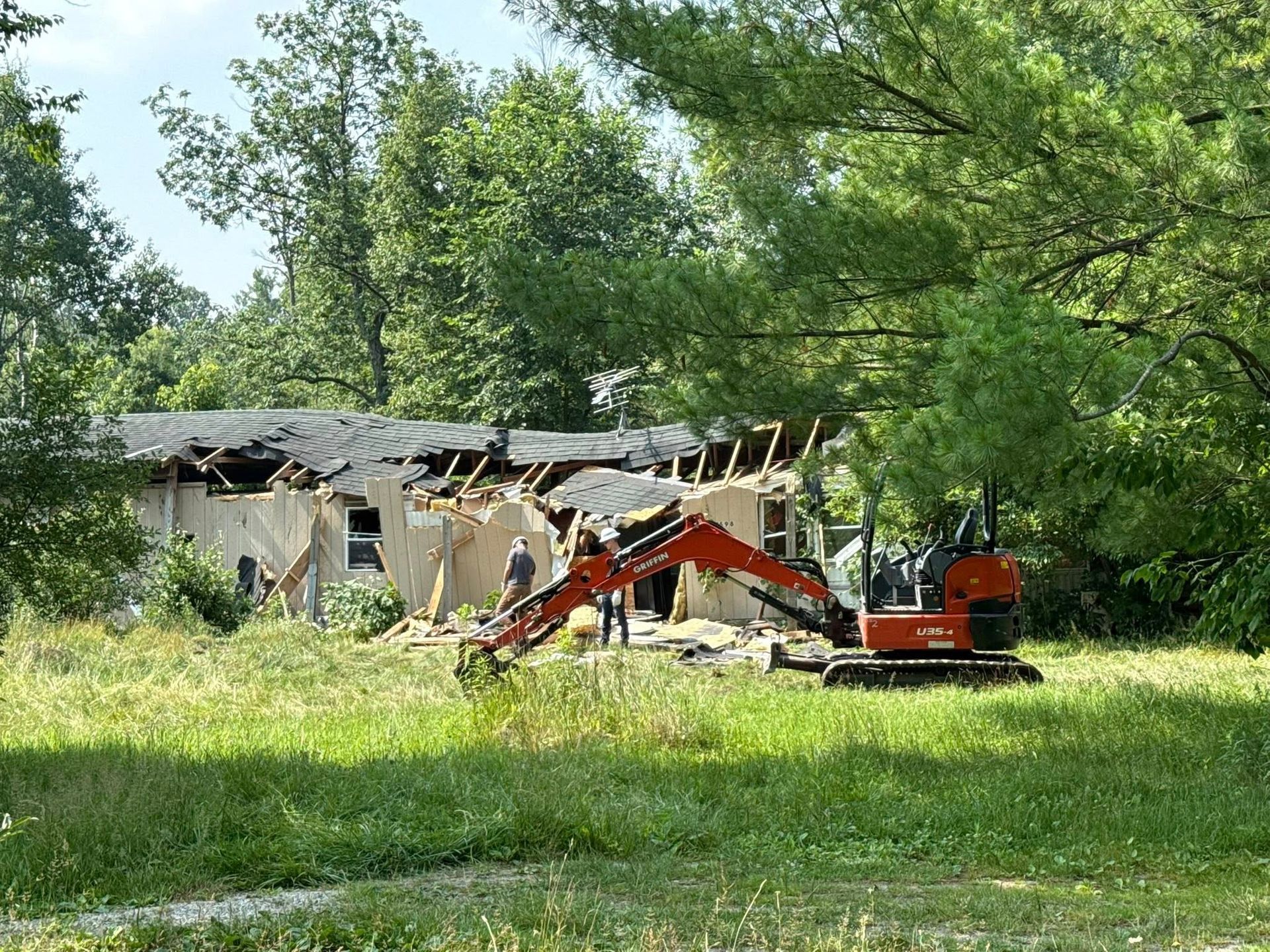 Mini excavator demolishing a house in a grassy field, trees in the background, two workers observing.
