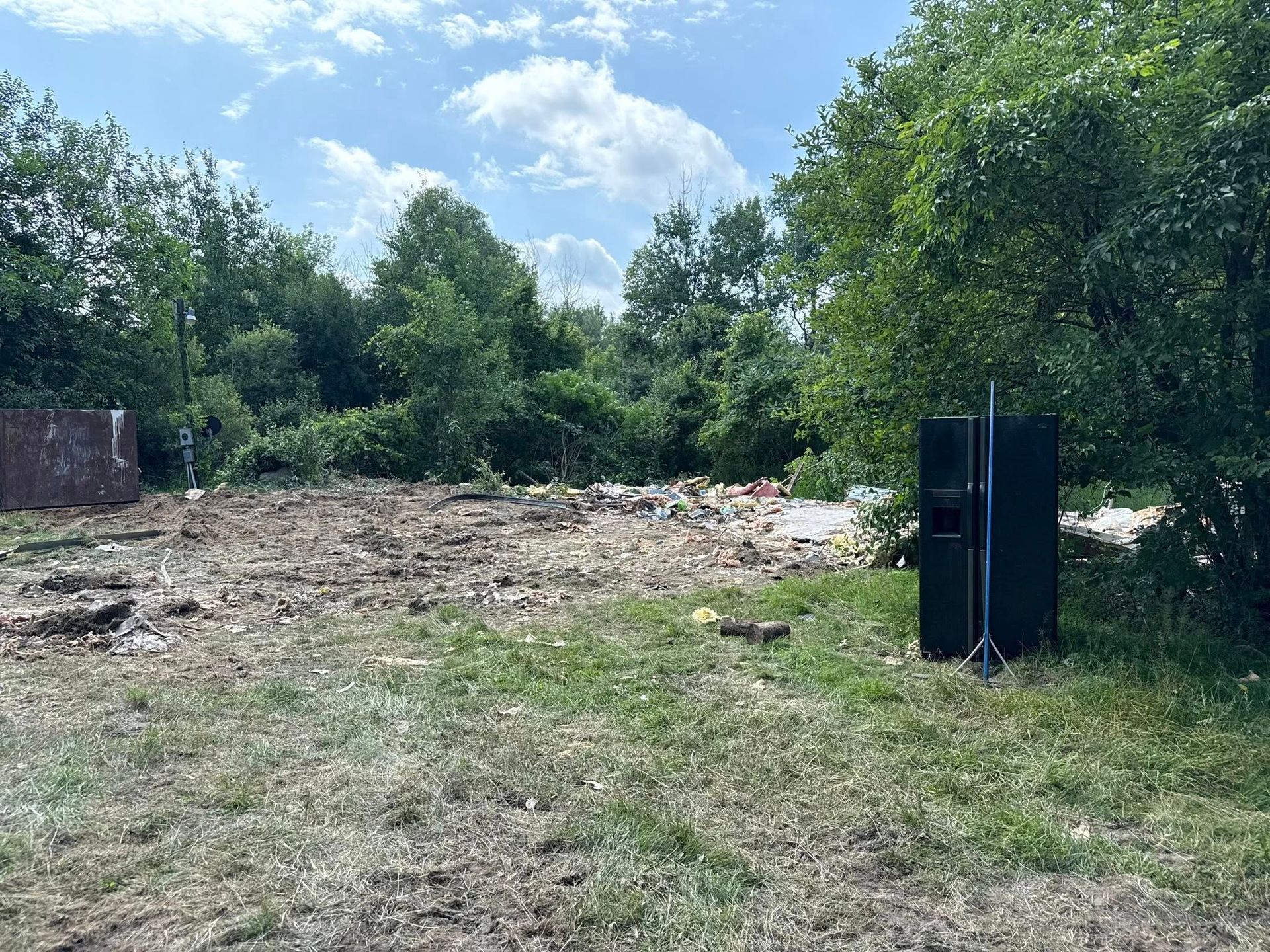 Grassy field with debris, trees, and a black speaker on a stand. Overcast sky.