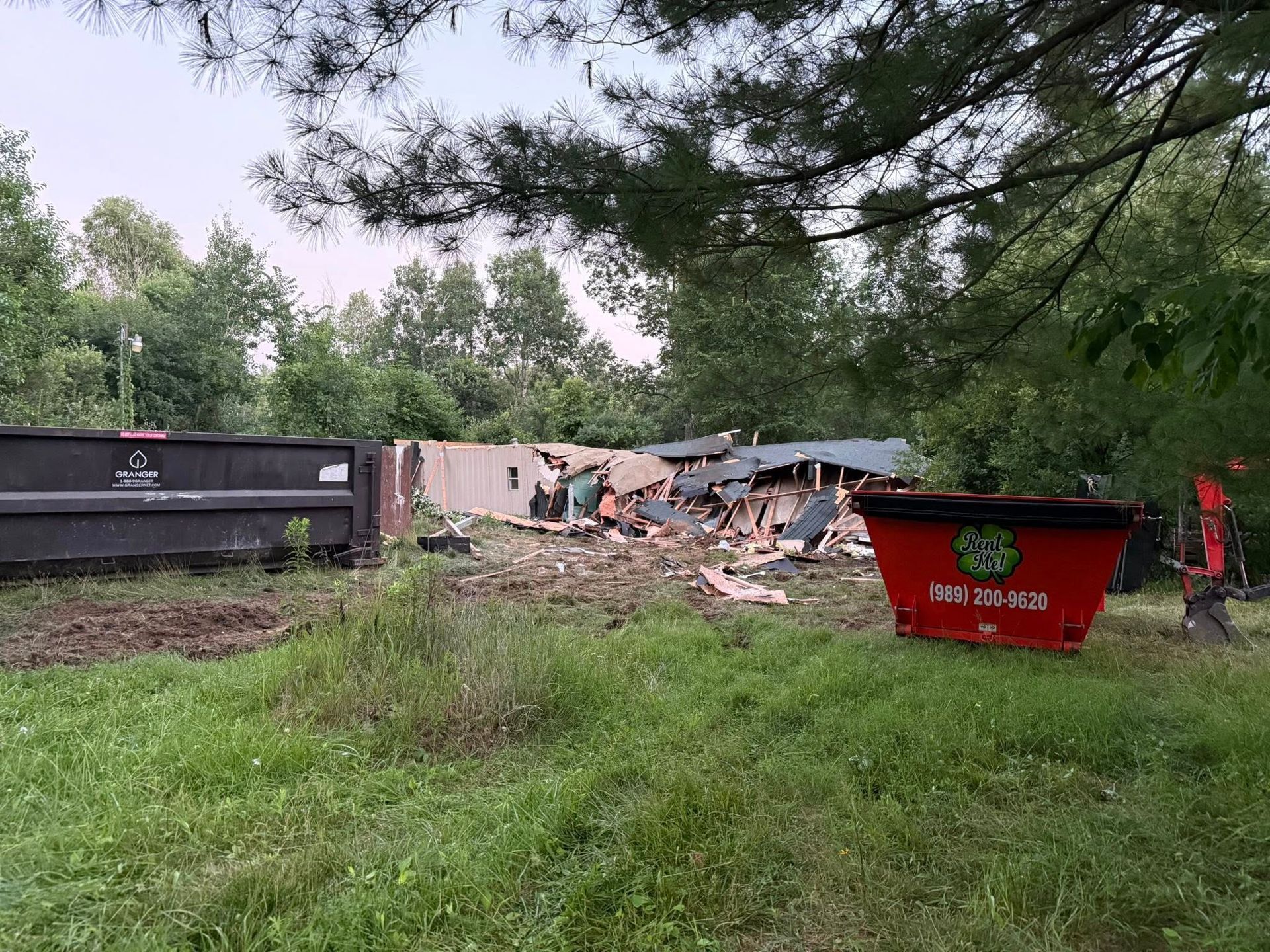 Demolished house with debris, two dumpsters on grassy property, trees in the background.