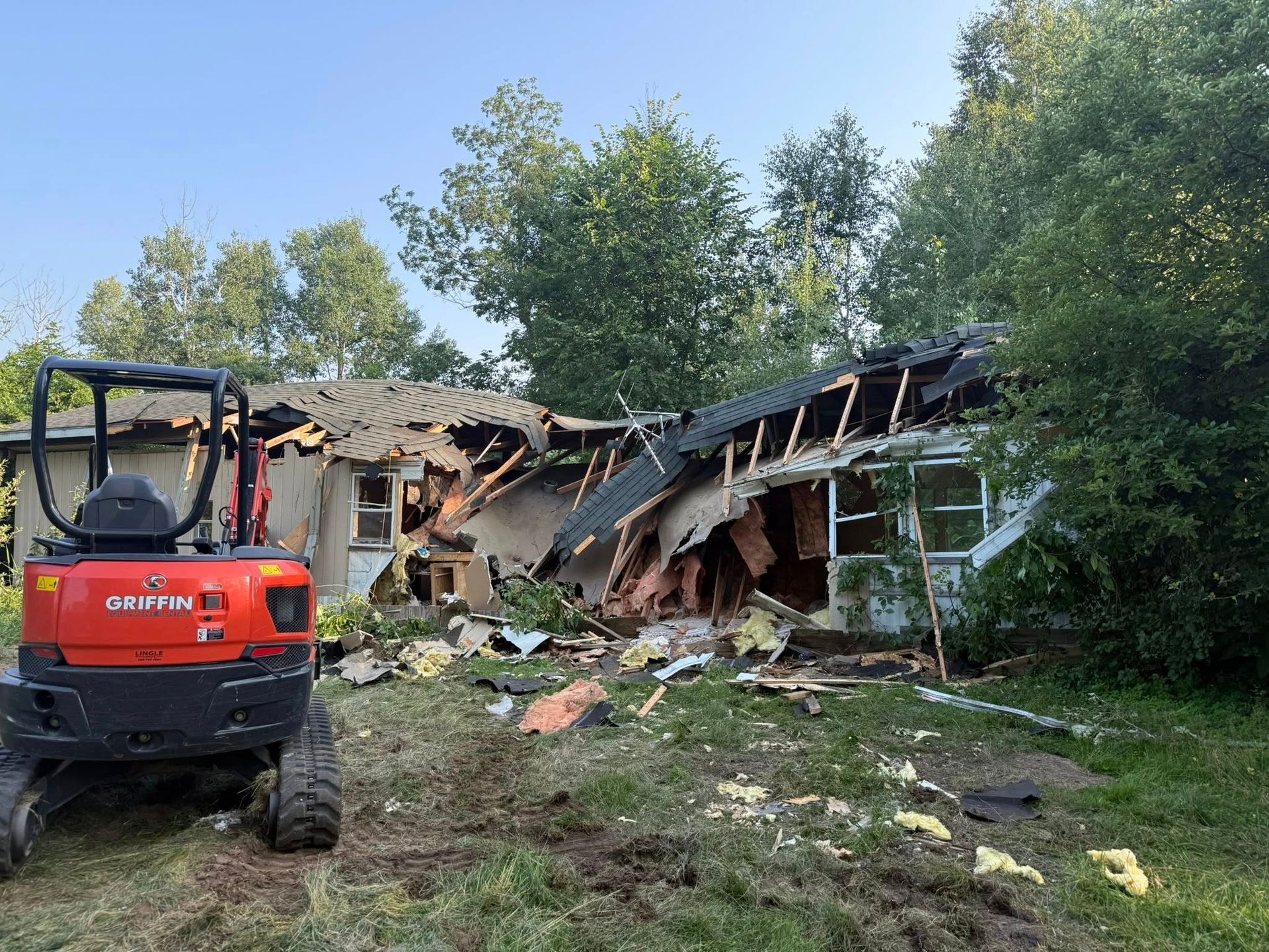 Demolished house with collapsed roof and orange excavator in yard. Trees and blue sky in background.