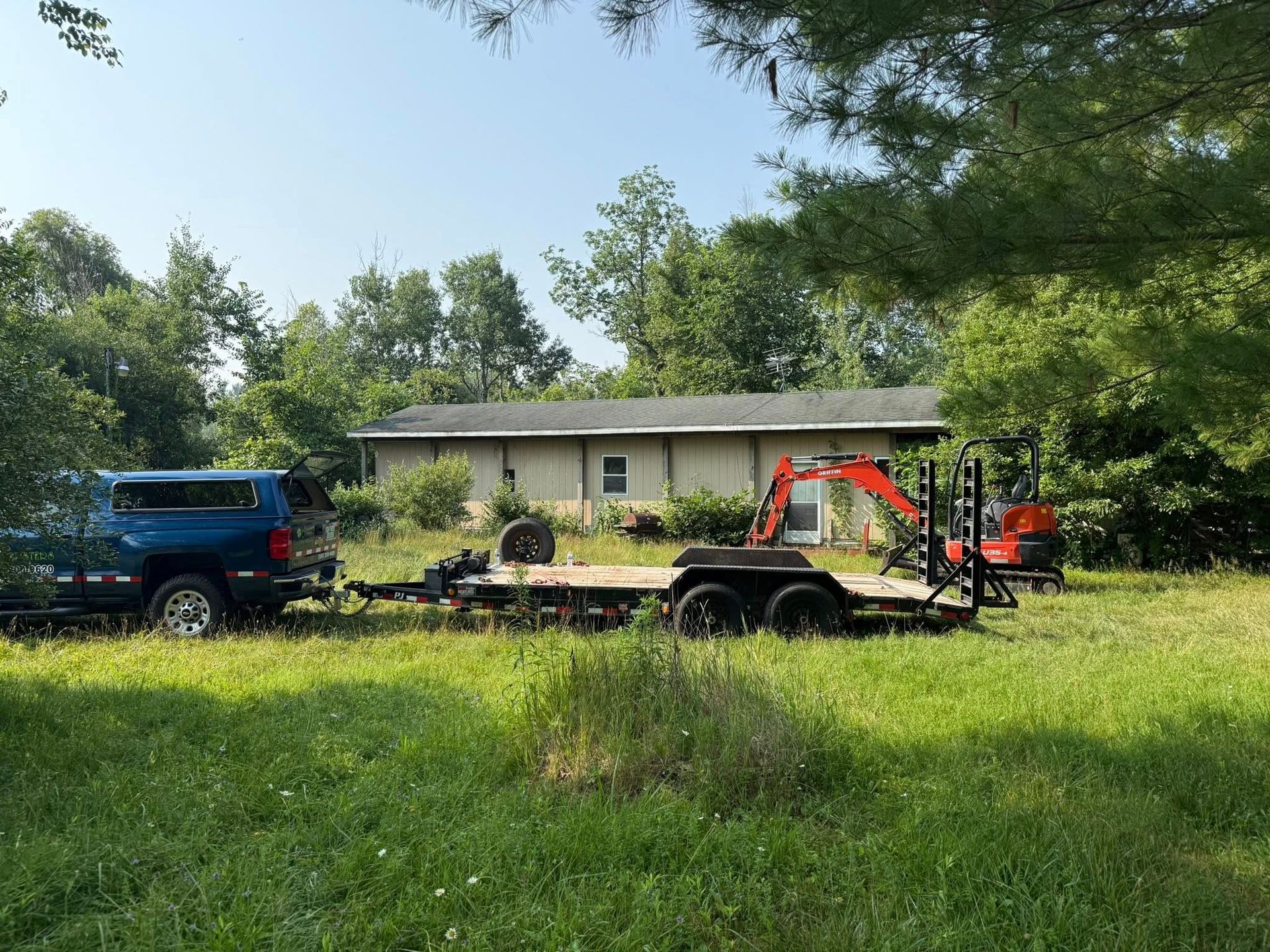 Blue truck towing a trailer with a small excavator parked in front of a beige house on a grassy field.