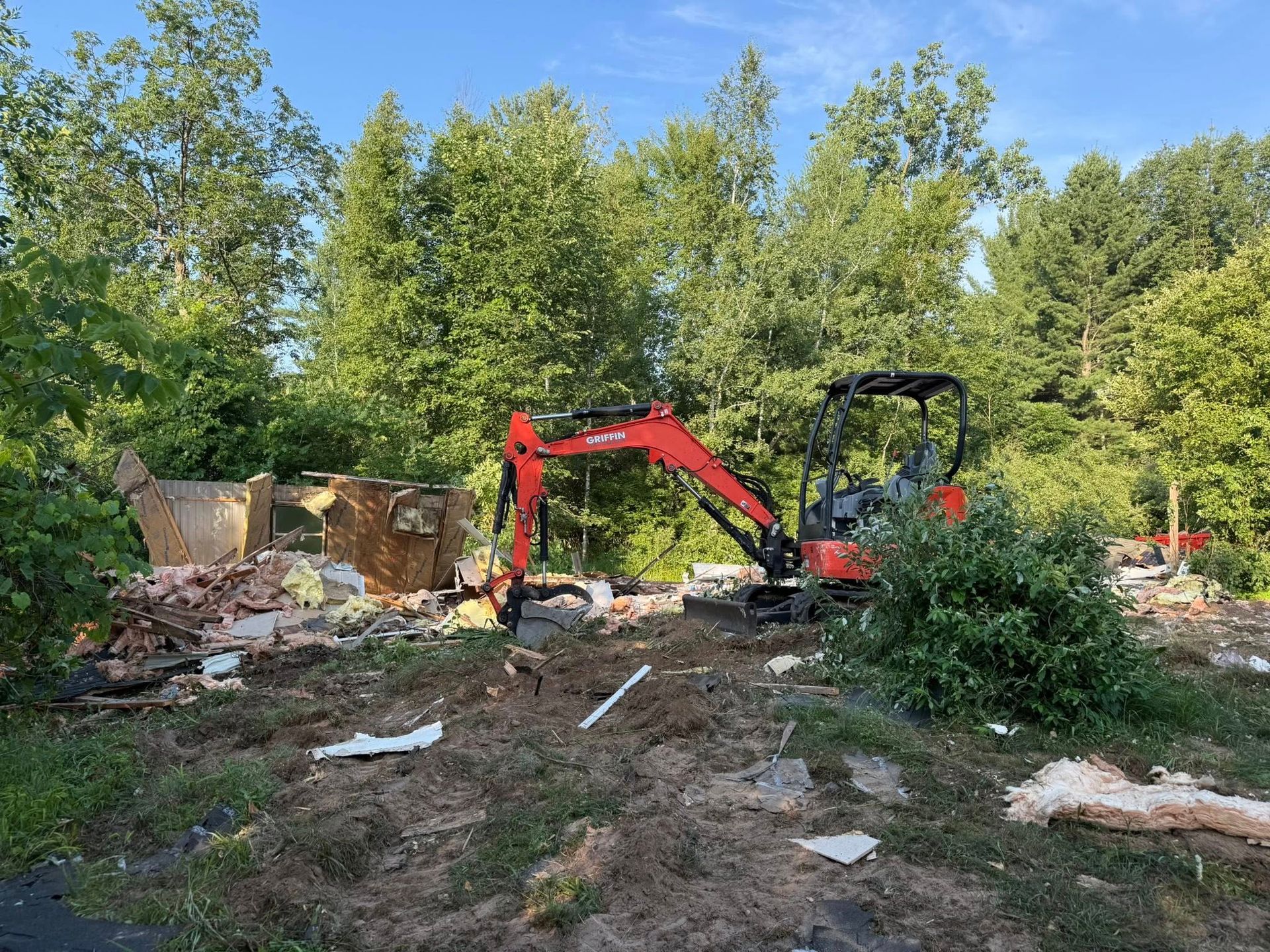 Orange excavator demolishing a structure, debris scattered on ground. Green trees in the background under a blue sky.