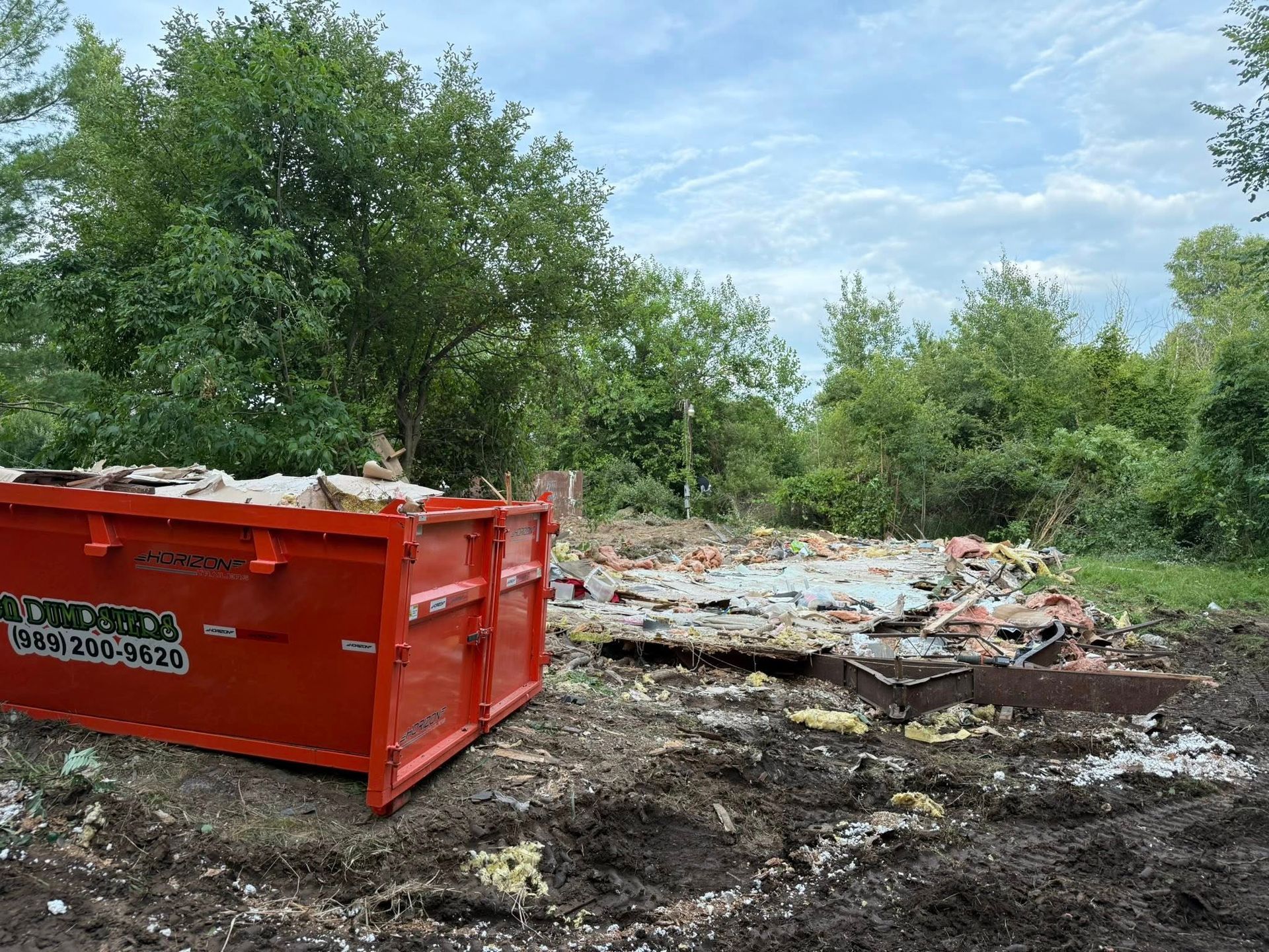 Red dumpster filled with debris next to a demolished building foundation in an overgrown lot.