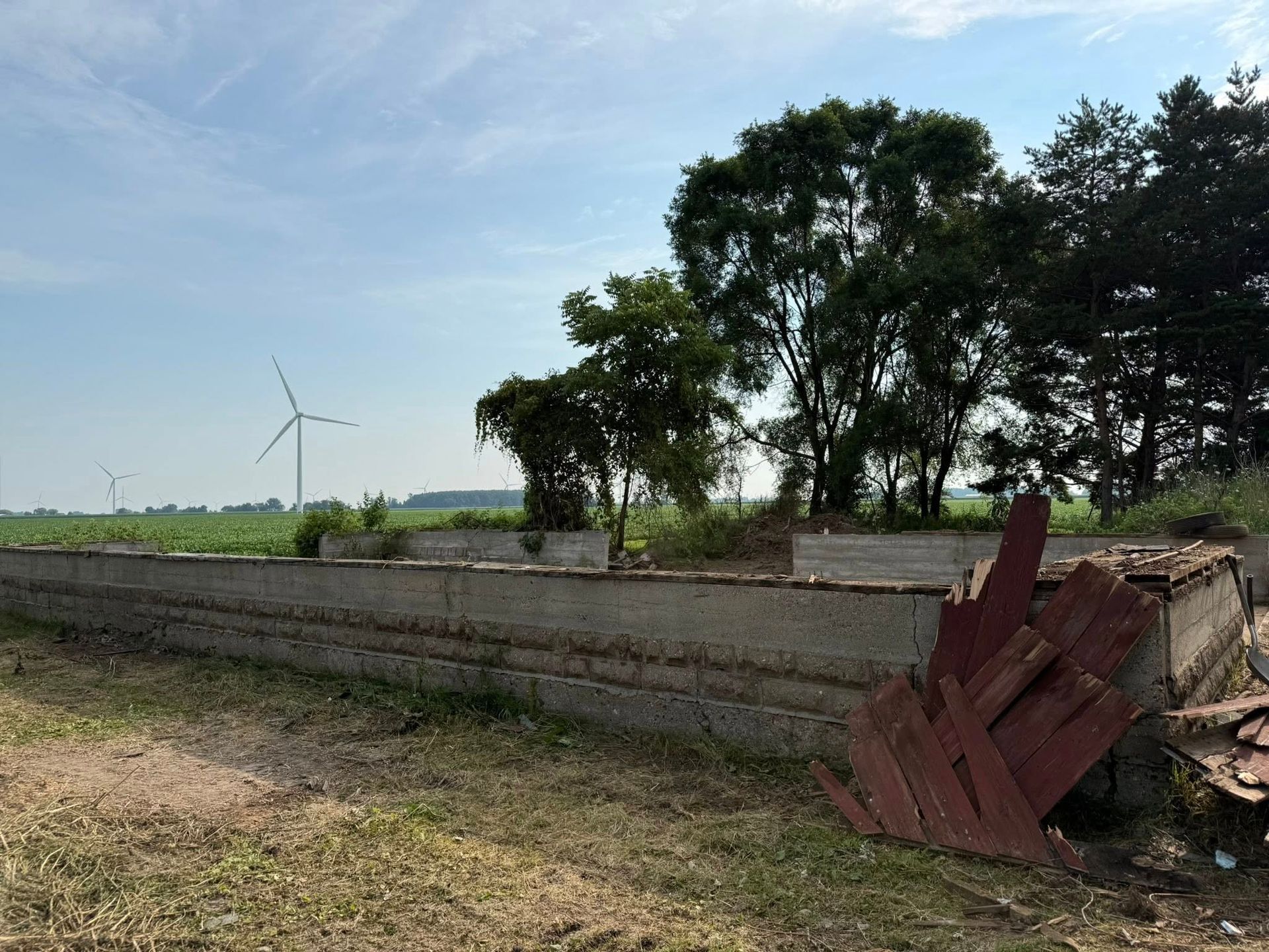 Low concrete wall with partial wood structure, field, trees, and windmill under a blue sky.