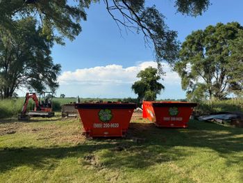 Two orange dumpsters on grass, with an excavator, trees, and blue sky.