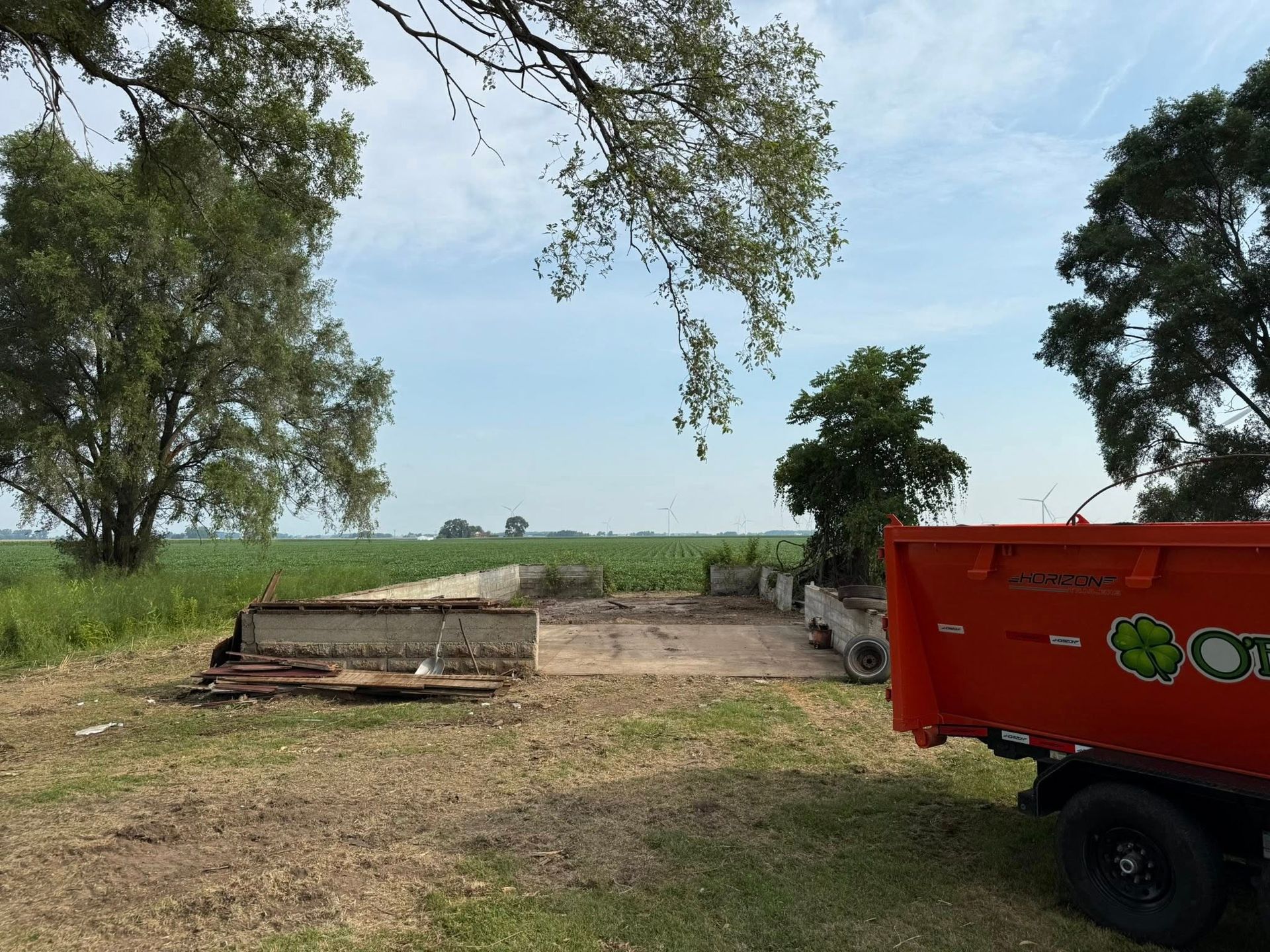 An empty concrete foundation, orange dump truck, and trees in a grassy field with a blue sky.