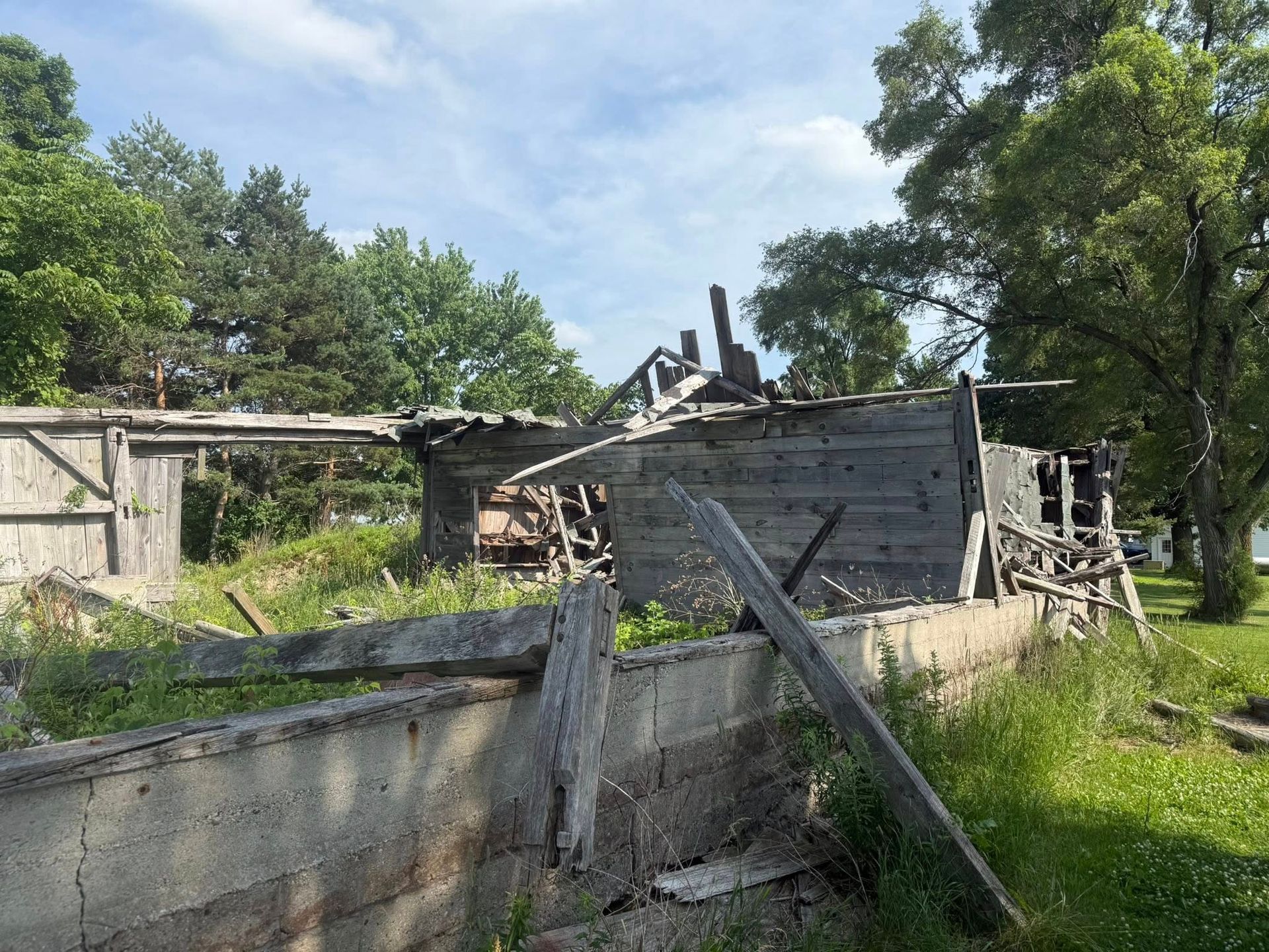 Dilapidated wooden structure with a collapsed roof, surrounded by grass and trees.