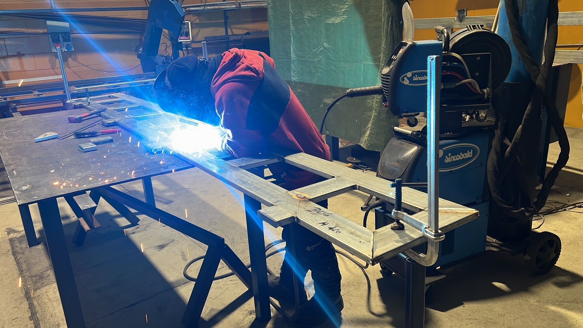 Un saldatore con la giacca rossa produce scintille su un banco da lavoro in un'officina.