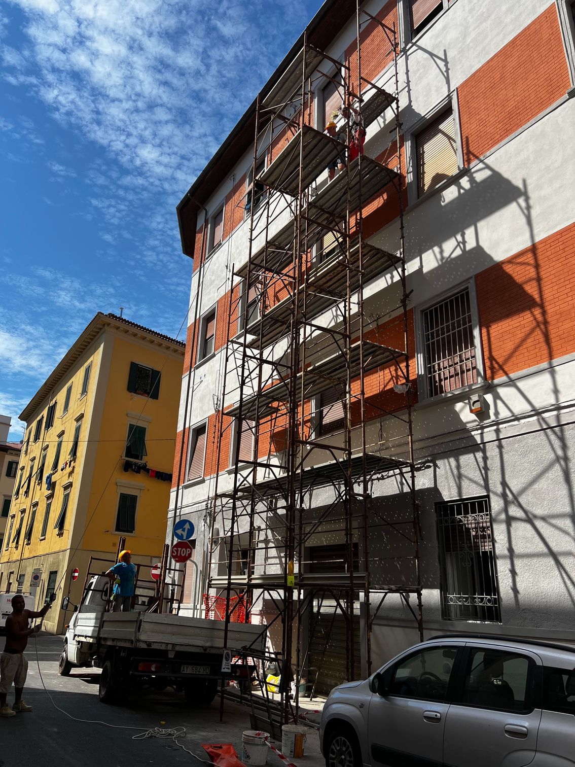 Palazzo di appartamenti con impalcature, facciata arancione e bianca, su una strada soleggiata, con un camion e un'auto parcheggiati sotto.