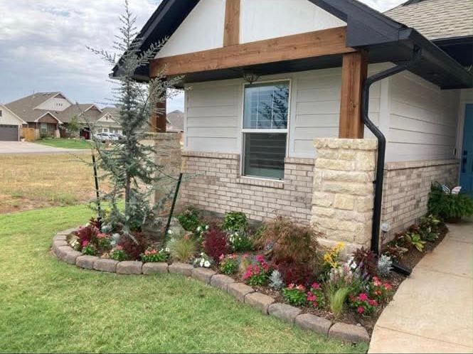 A tan brick house exterior with stone pillars and a landscaping bed filled with a small evergreen and colorful flowers.