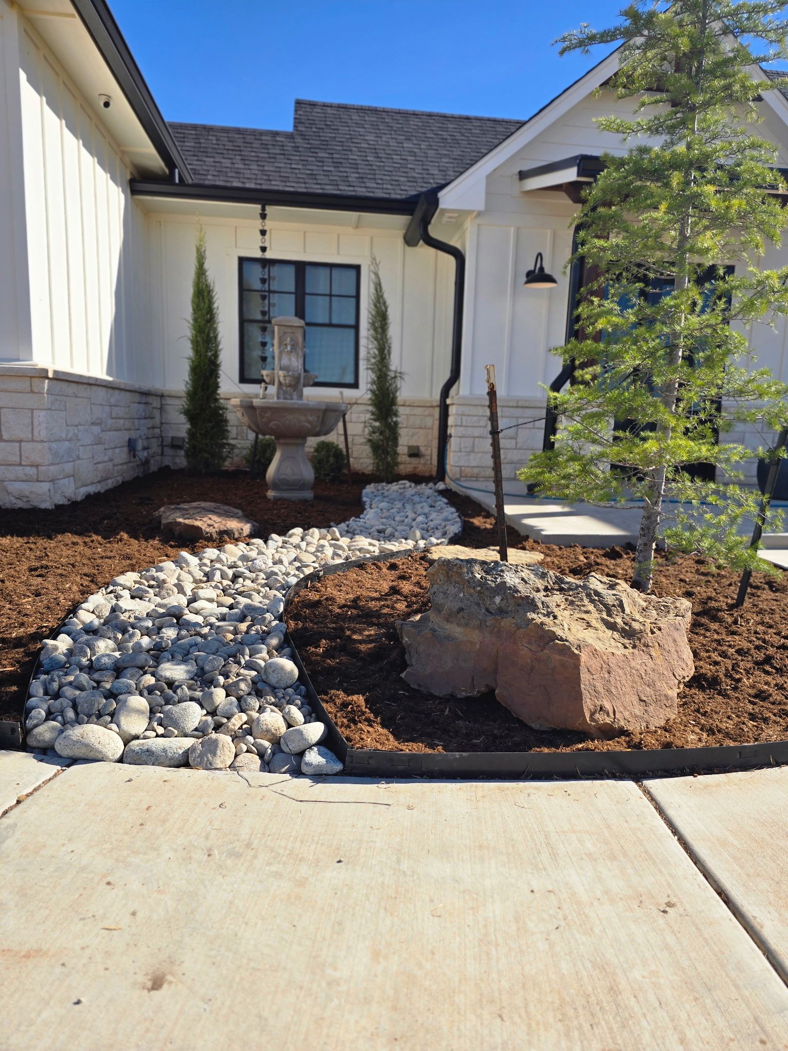 A stone path winds through mulch in a landscaped front yard with a fountain near a white modern farmhouse-style house.