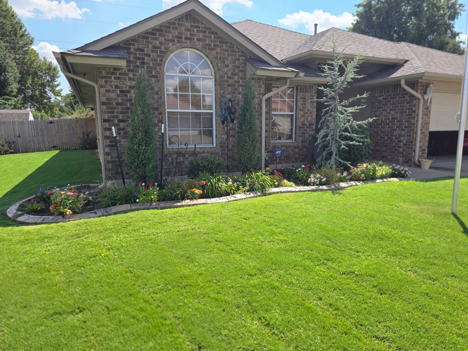 A brown brick house with a front garden featuring shrubs, roses, and a curved stone border under a bright blue sky.