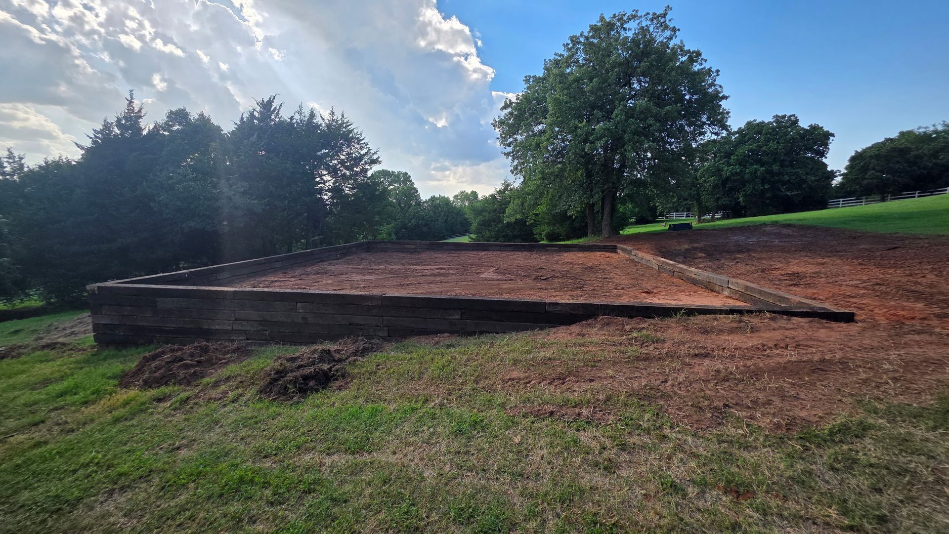 A low, rectangular retaining wall made of dark wooden beams surrounds a patch of raised, reddish-brown soil in a field.