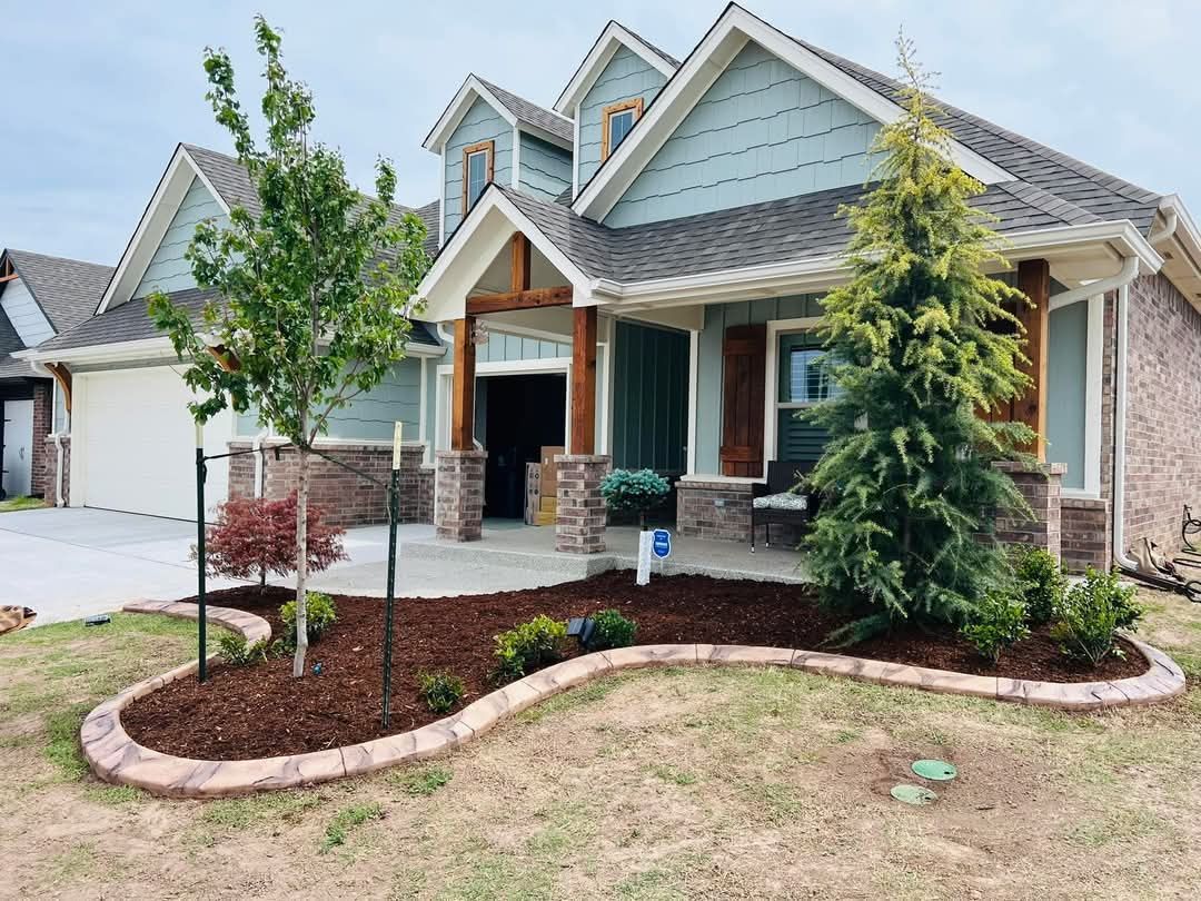 A suburban house with light blue siding, brick base, and a new landscaped garden bed with mulch and young trees.
