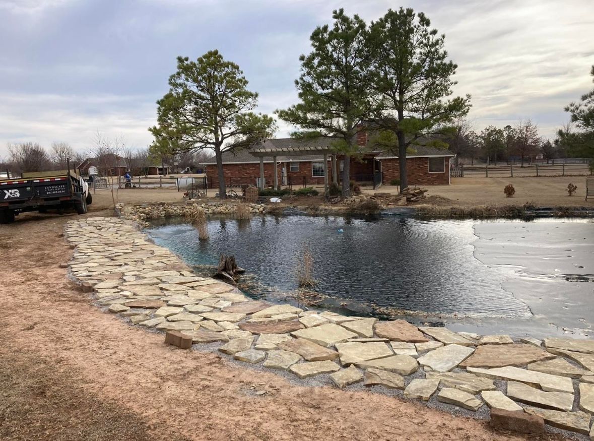A stone patio borders a calm pond in a rural setting, with a brick building and trees in the background under a cloudy sky.