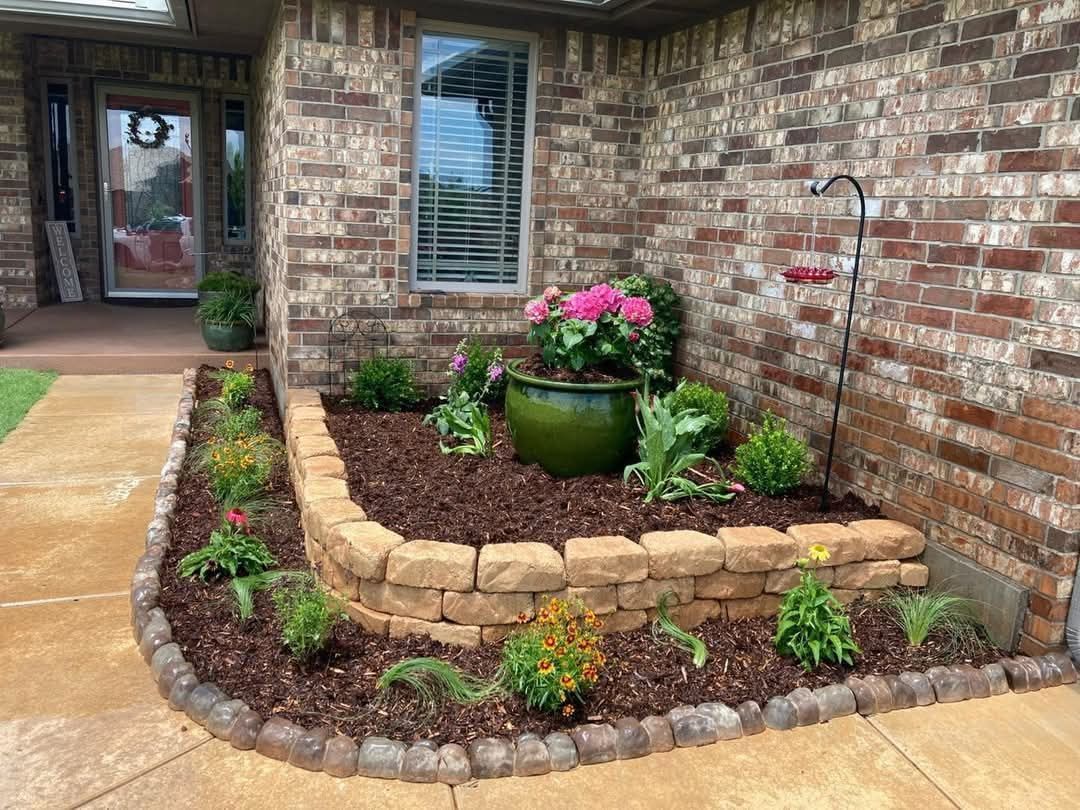 A tiered brick garden bed with a large green pot, various plants, and brown mulch next to a brick house and walkway.