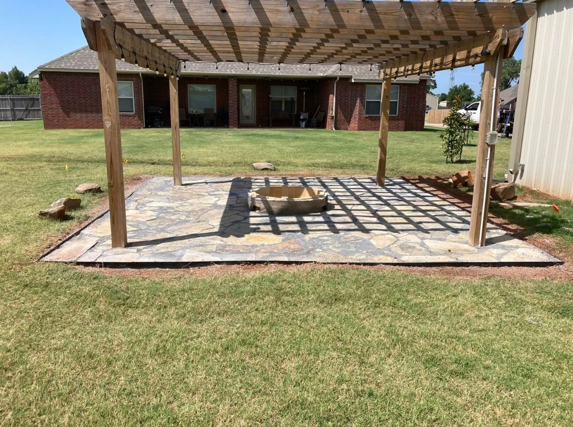A wooden pergola stands over a stone patio with a central fire pit in a residential backyard.